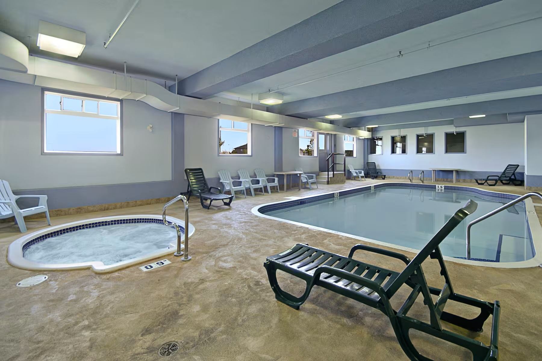 Indoor pool area with hot tub, lounge chairs, tables, windows, exposed beams, and calm, well‑kept surroundings.