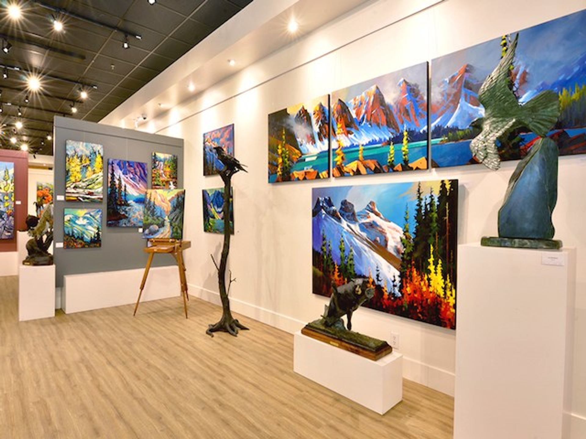 Gallery wall with bright mountain paintings and wildlife sculptures on white display stands.
