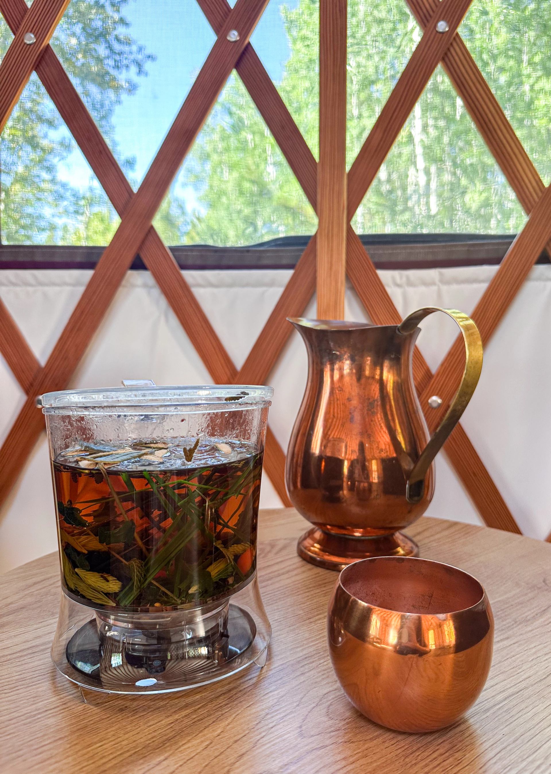 Herbal tea steeps beside a copper pitcher and cup on a wooden table.