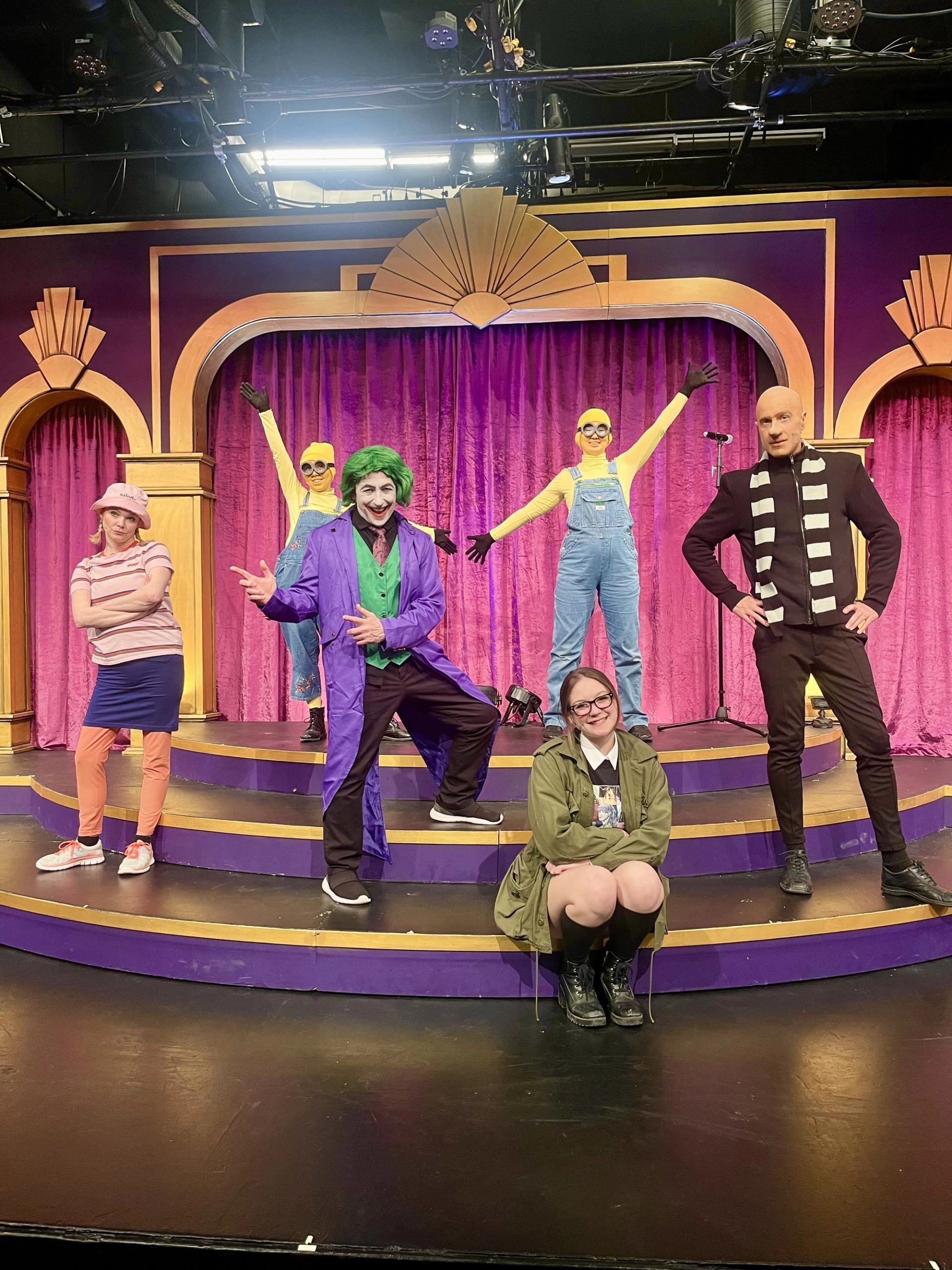 Costumed performers pose on a purple and gold theatre stage.