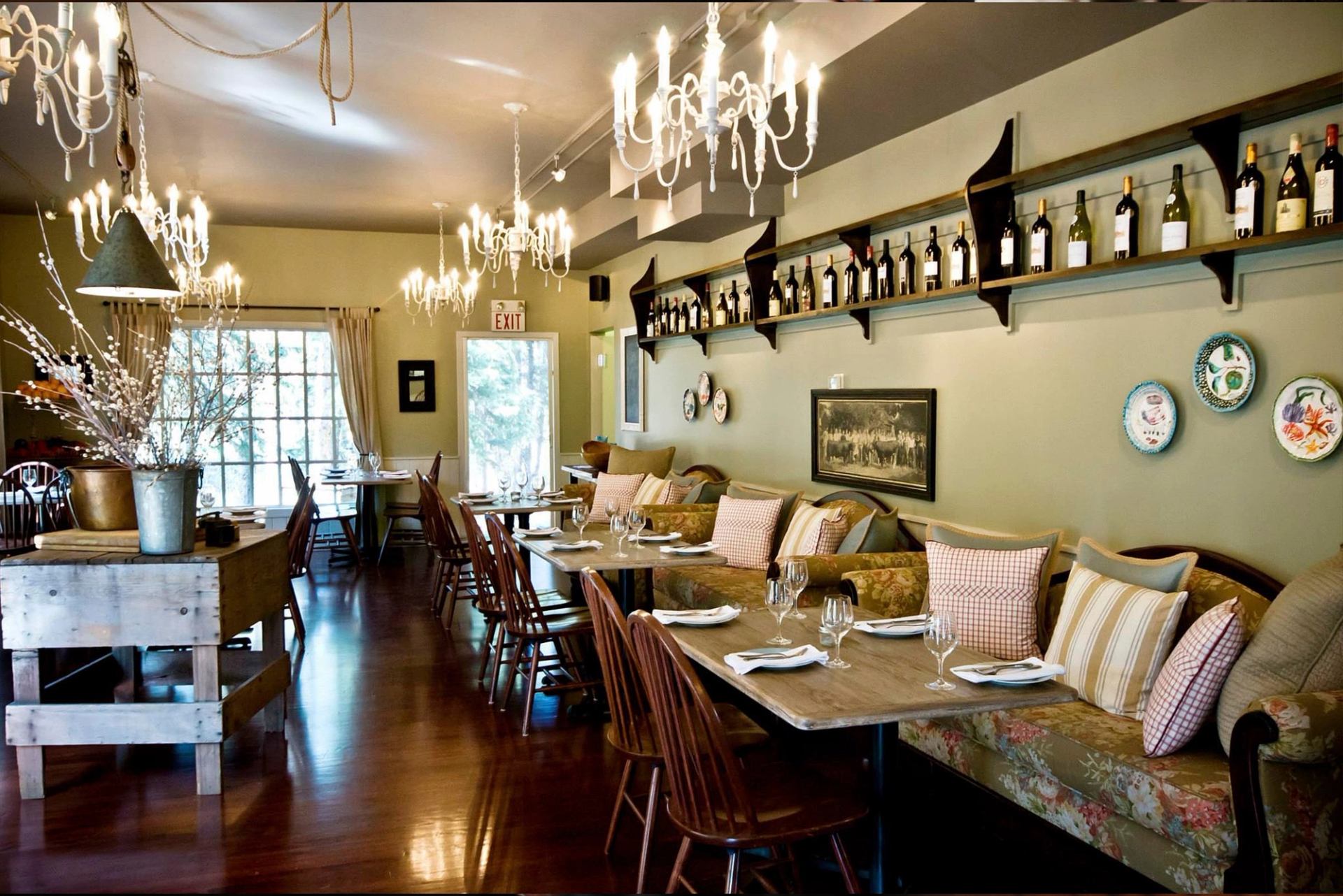 Restaurant dining room with set tables, chandeliers, cushioned benches and wine bottles.