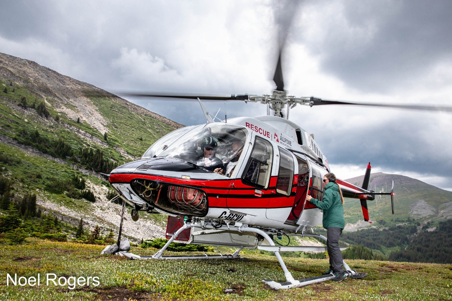 Rescue helicopter on alpine meadow as person loads gear, mountains and clouds in background.