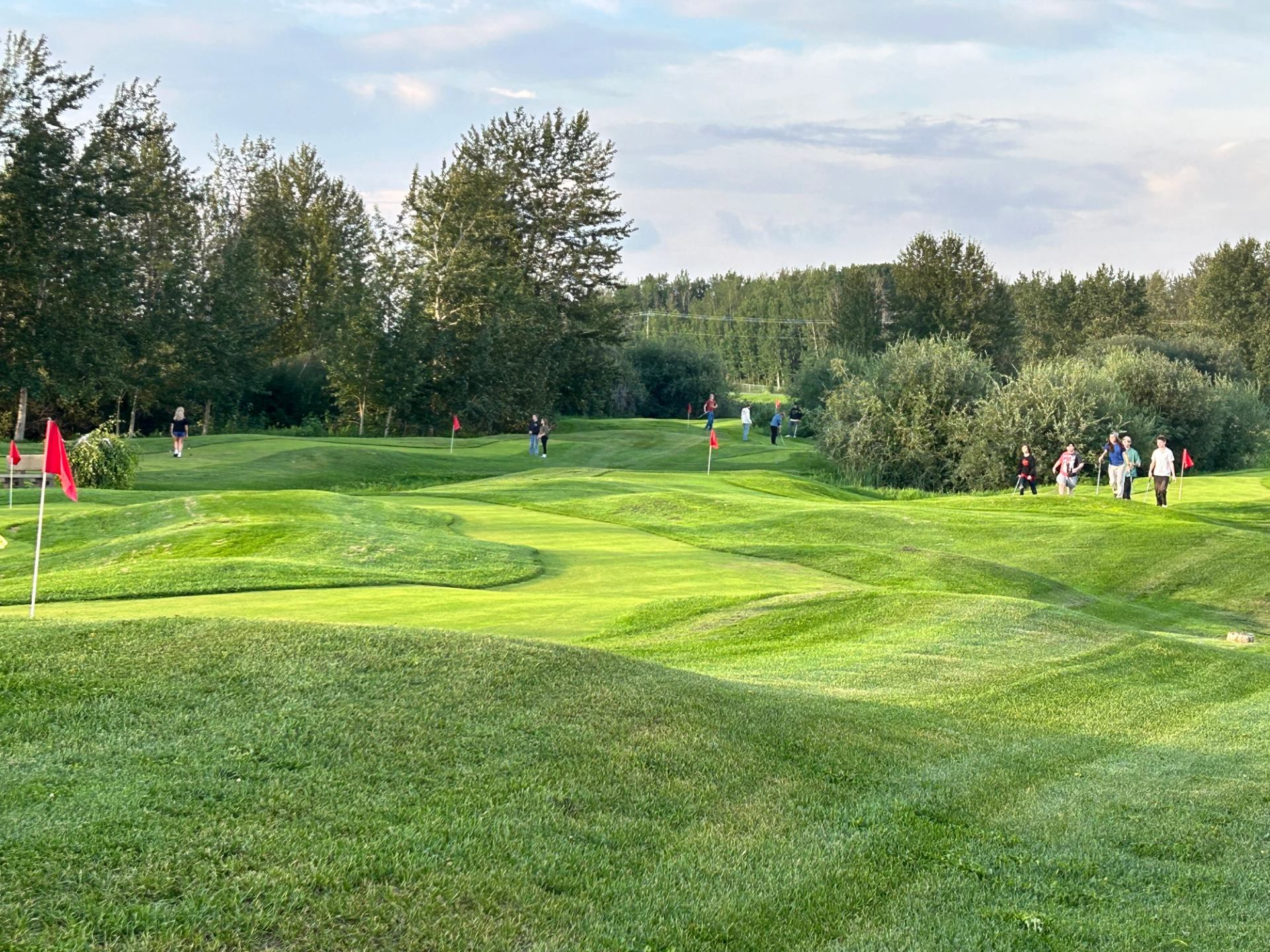 People walking and playing on a green, hilly putting course with red flags.