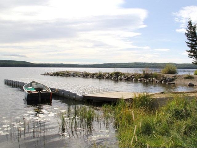 Small boat at a lakeside dock with calm water and a rocky shoreline.
