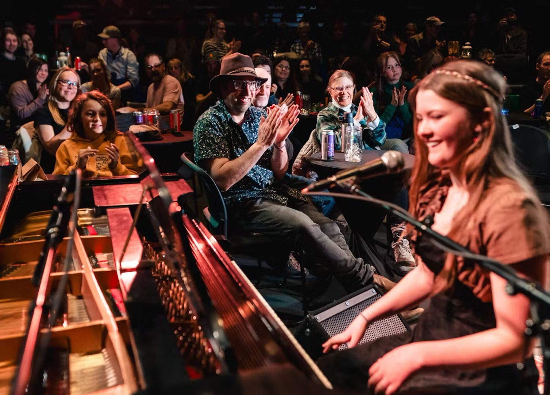 Performer at piano with mic during open mic night, audience clapping in cozy venue.