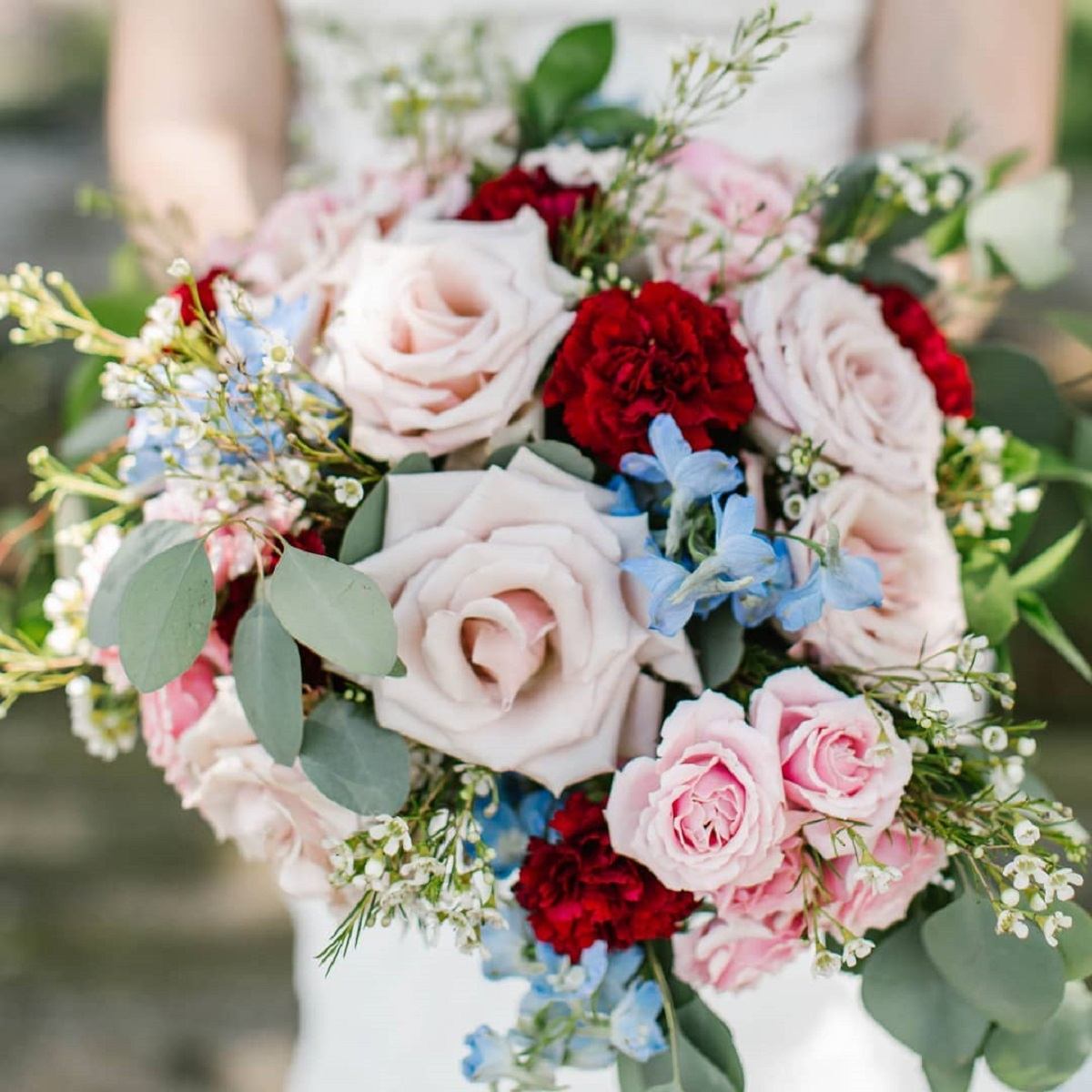 Hand holding bouquet with roses, carnations, delphiniums, and greenery.