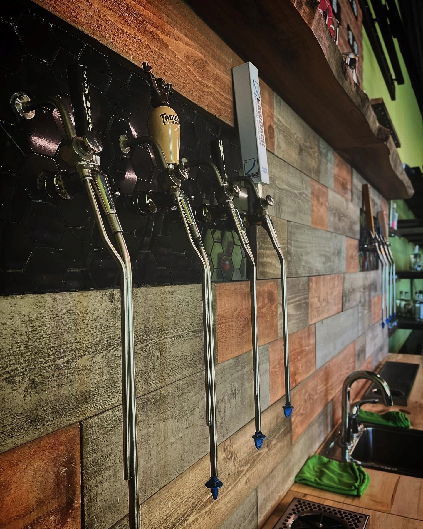 Row of beer taps on wooden wall with green trays below in a rustic bar setting.