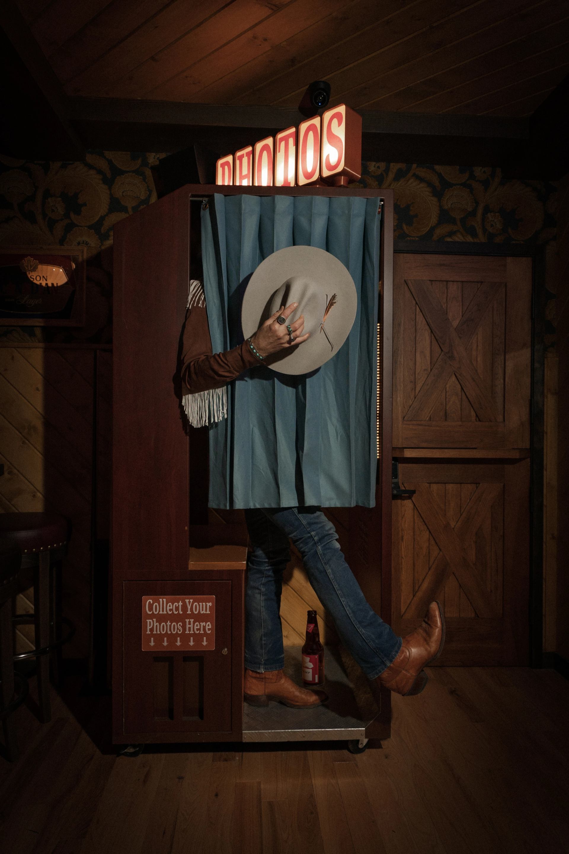 Person in a photo booth holding a cowboy hat, one boot stepping out onto the floor.