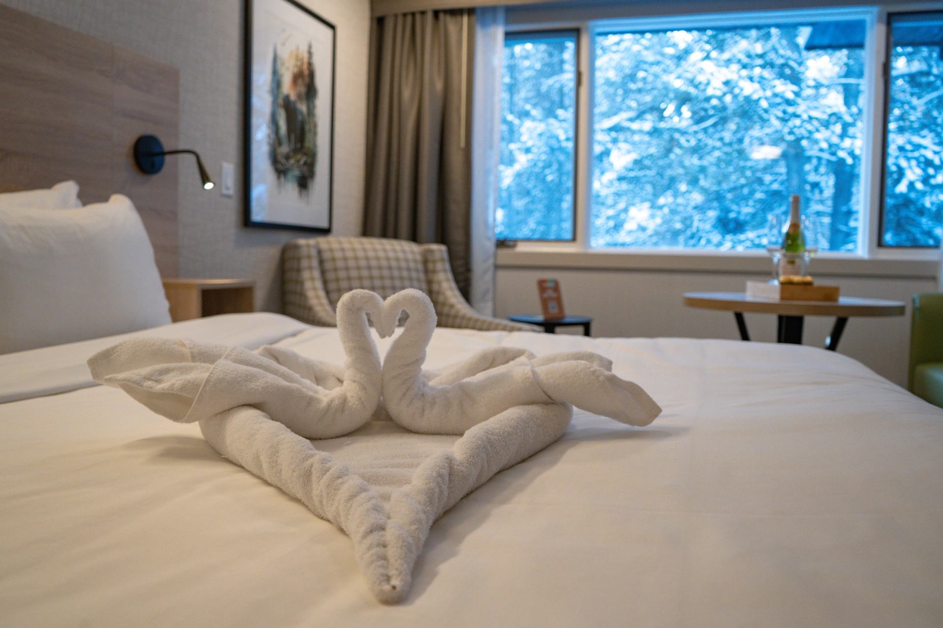 Towel swans arranged on a hotel bed with a snowy window view and seating area in the background.