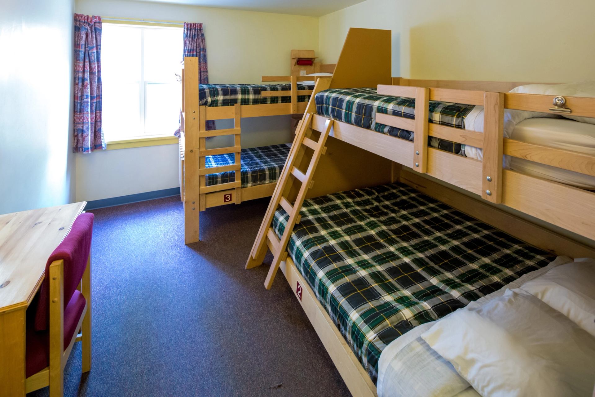 Four-bed dorm room at HI Banff with wooden bunk beds, plaid bedding, and large window.