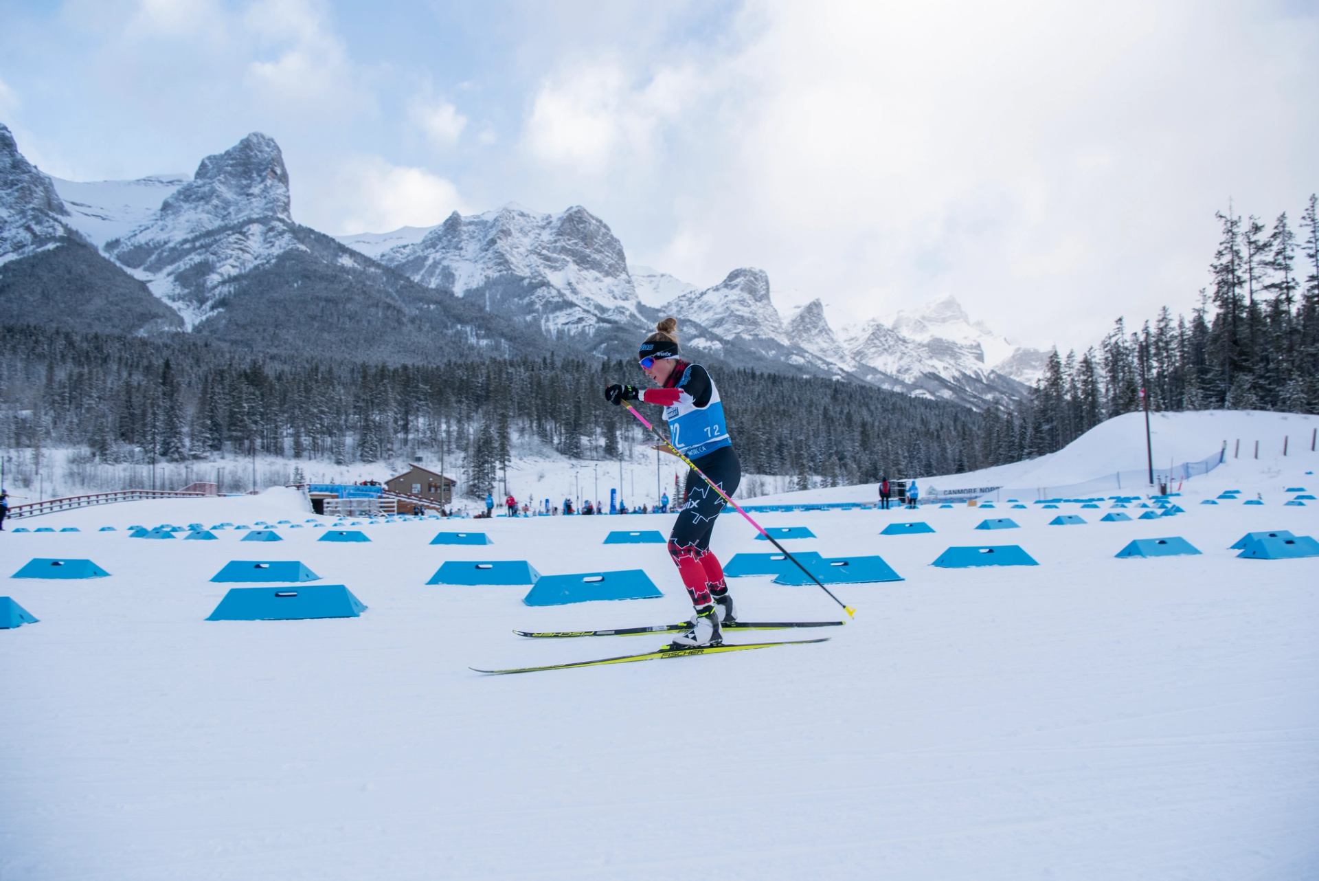 Para biathlete skiing past blue markers with snowy mountains behind.