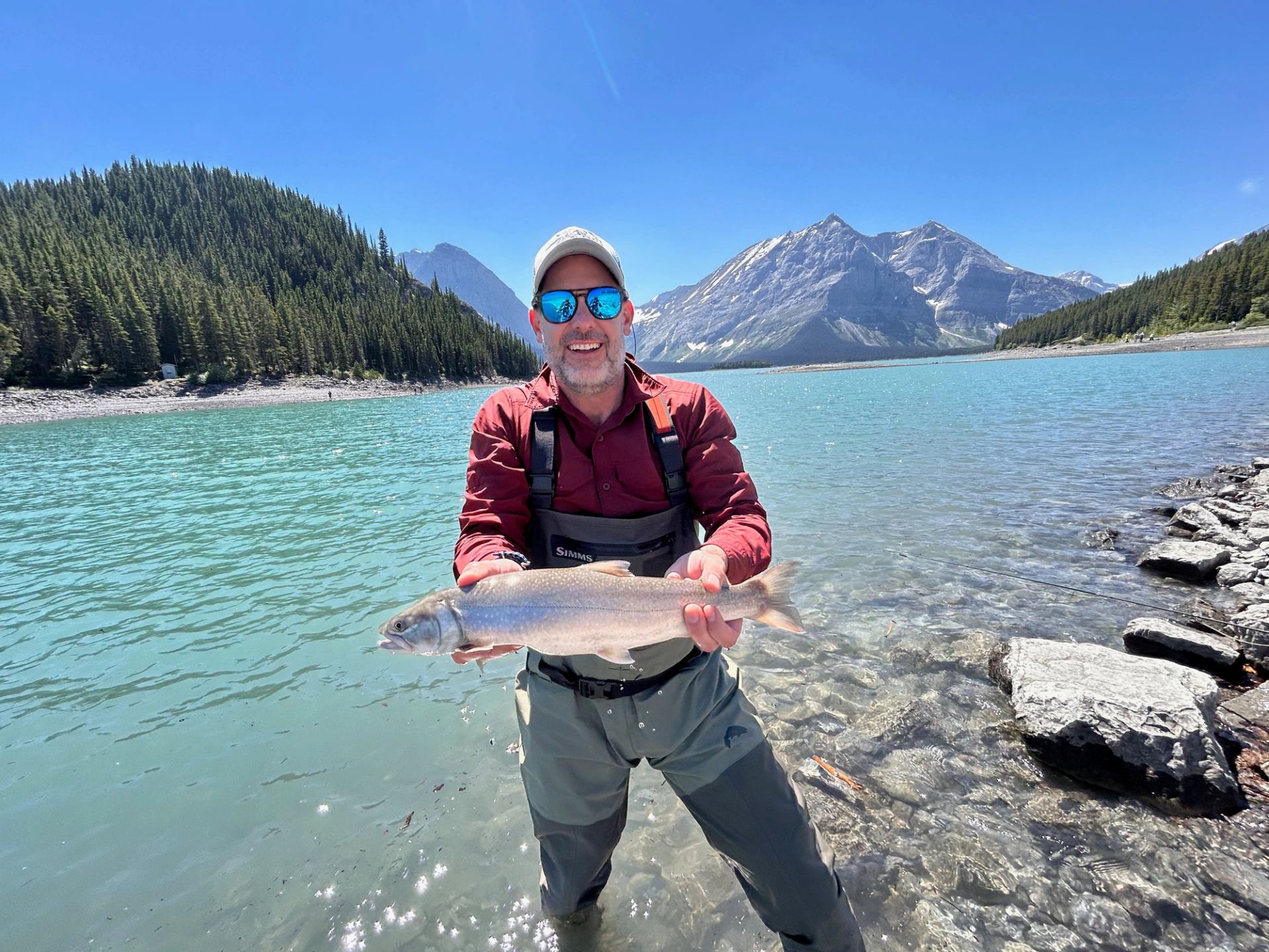 Angler standing in clear turquoise lake, holding fish with forested mountains behind.