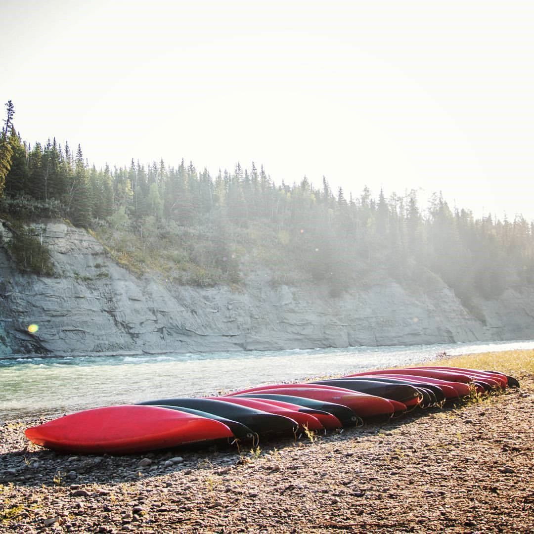 Red kayaks rest on a rocky shore beside a forested river at HeLa Ventures.