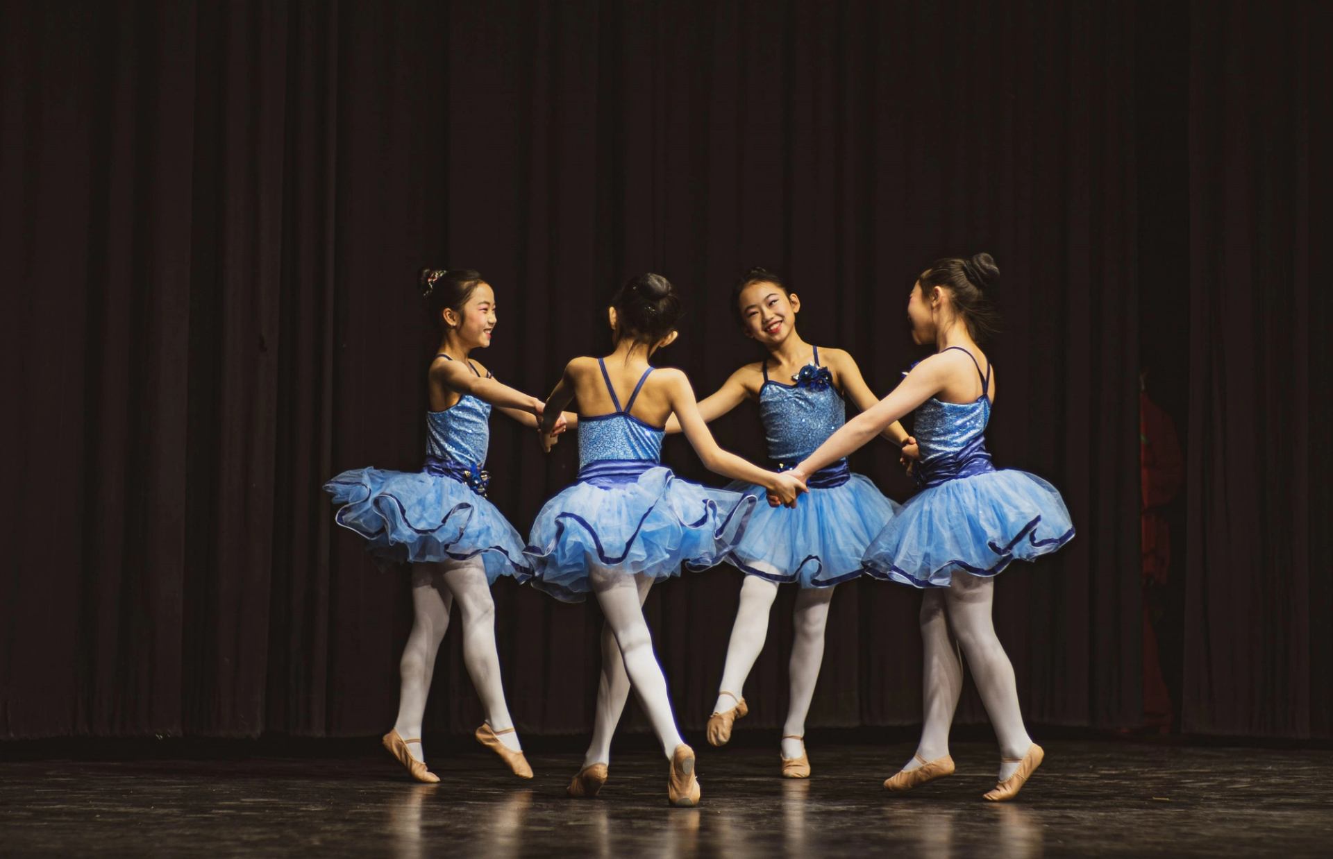 Four young dancers in blue tutus performing a synchronized routine on stage.