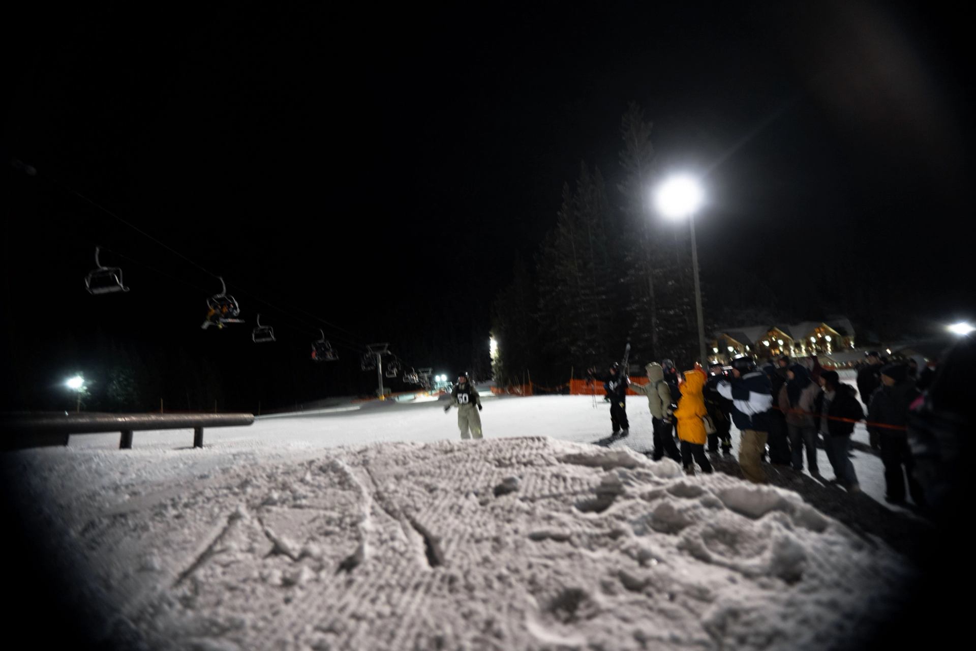 Crowd gathered at Backyard Rail Jam under bright lights with ski lift chairs in the background.