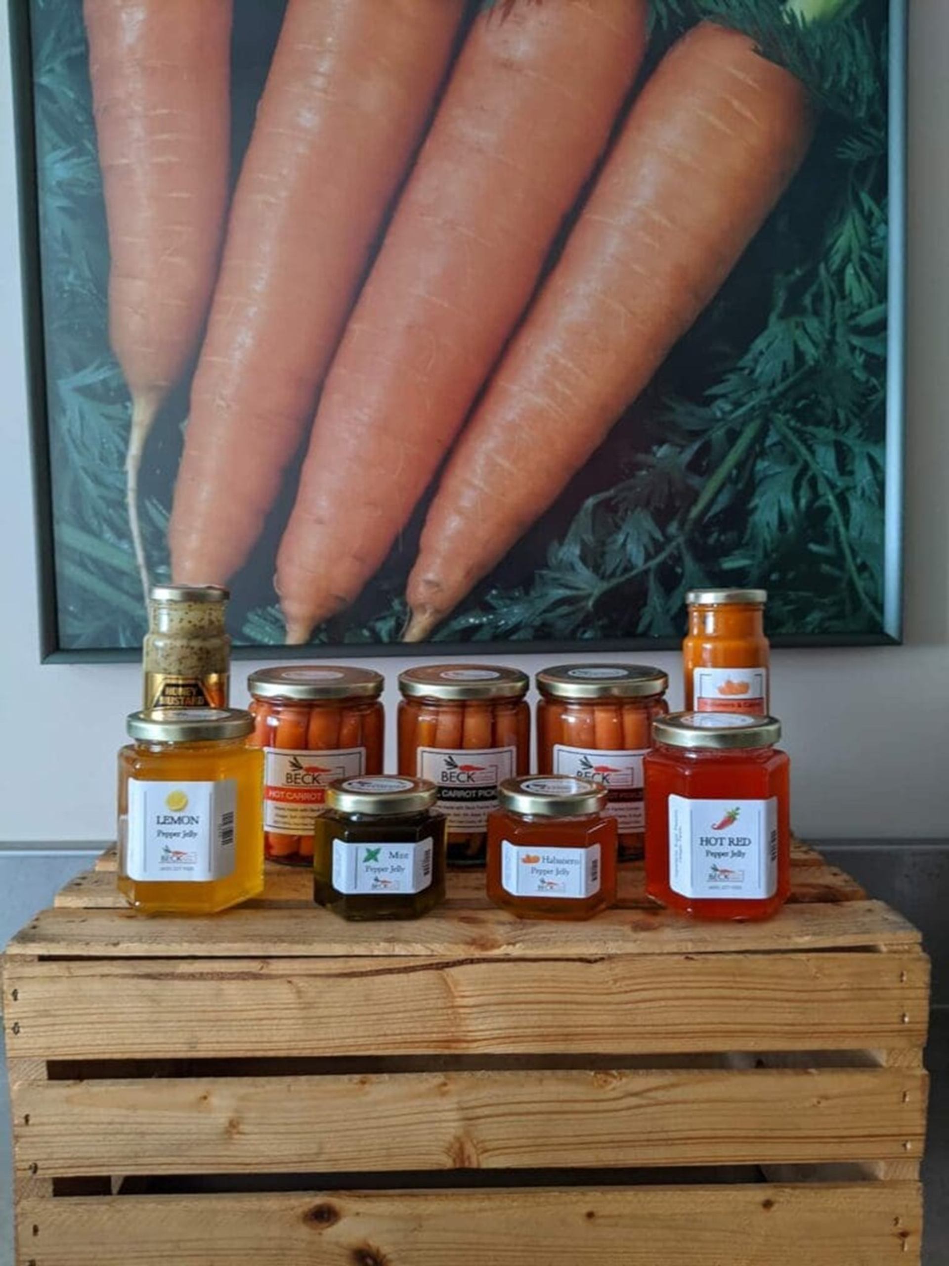 Wooden crate displaying assorted carrot pickles and pepper jellies before large carrot poster.