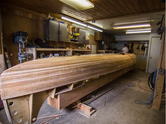 A workshop with wooden canoe construction in progress. A craftsman works on the canoe, surrounded by tools and shelves. The setting is warm and focused.