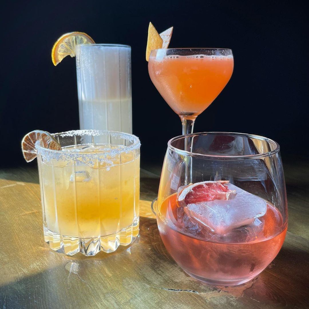 Four colorful cocktails with fruit garnishes on a wooden surface.