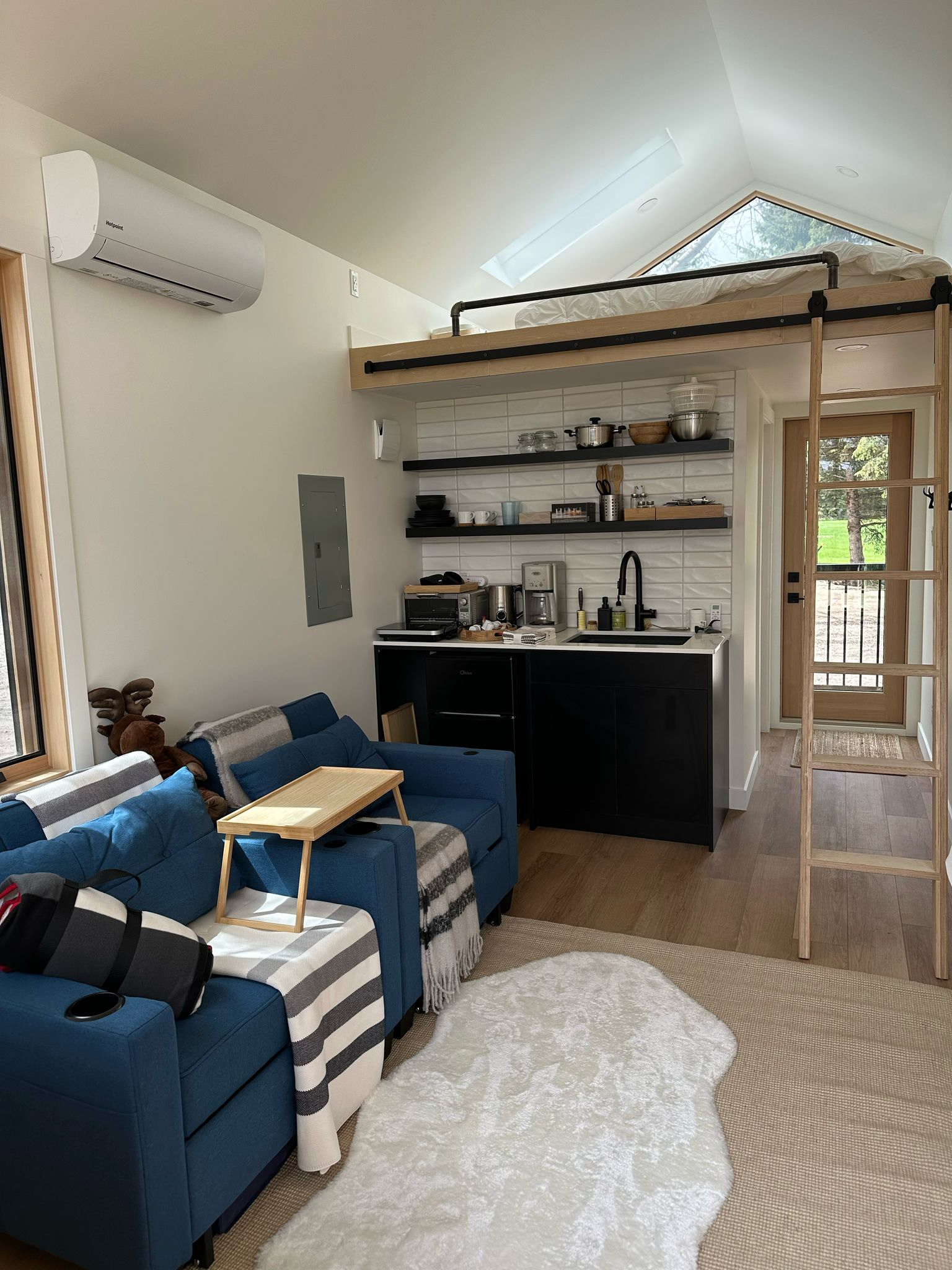 Interior of a small modern cabin with blue chairs, a kitchen area, and a loft bed under a skylight.