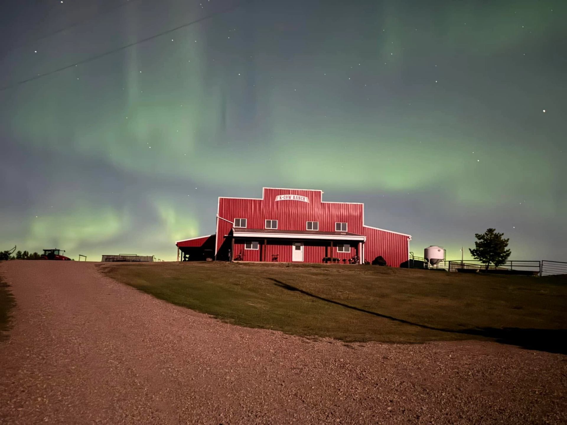 Red barn-style building under green aurora borealis, seen from gravel driveway at night.