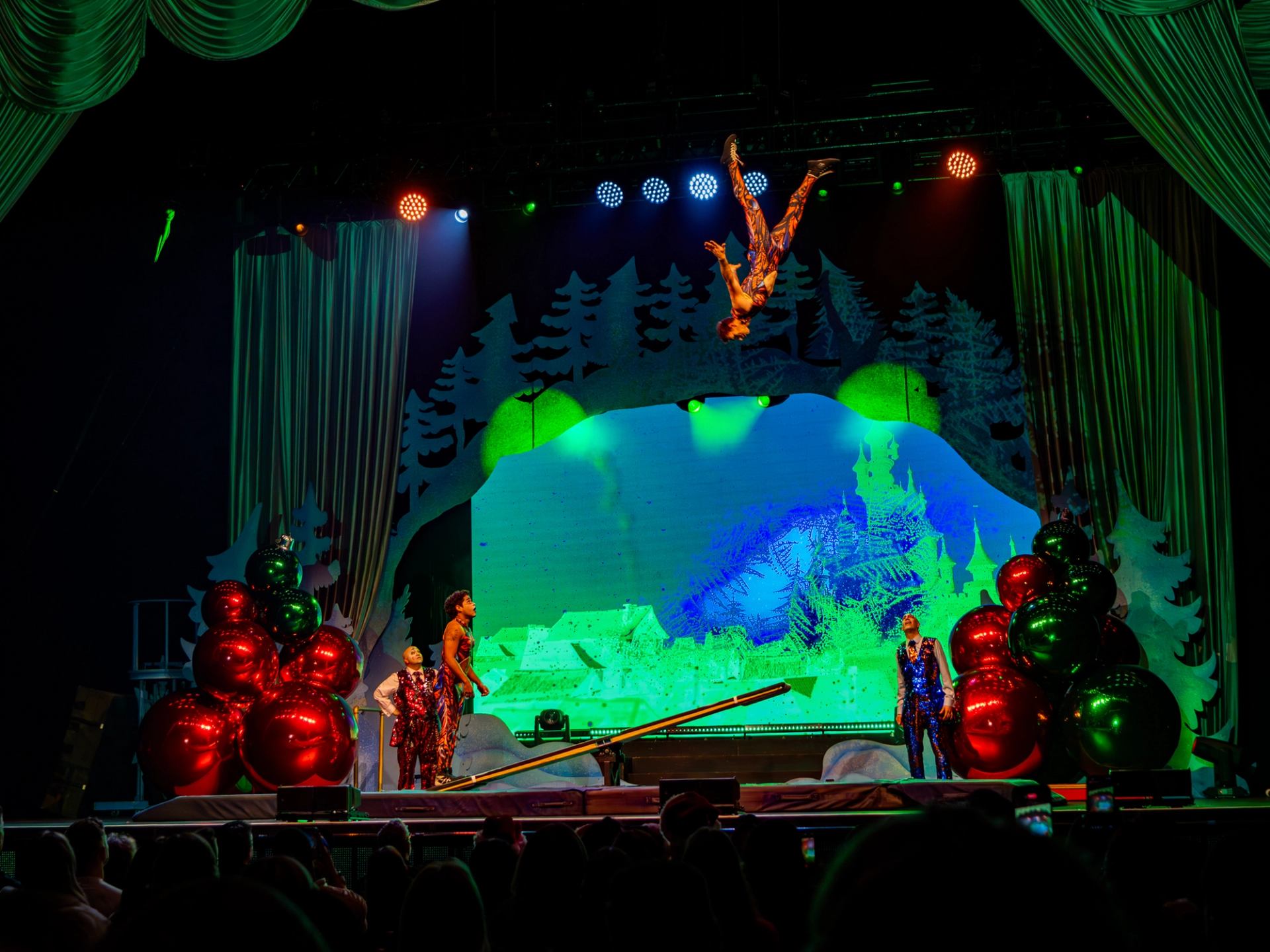 Acrobat flips above a seesaw as performers watch on a colourful theatre stage.