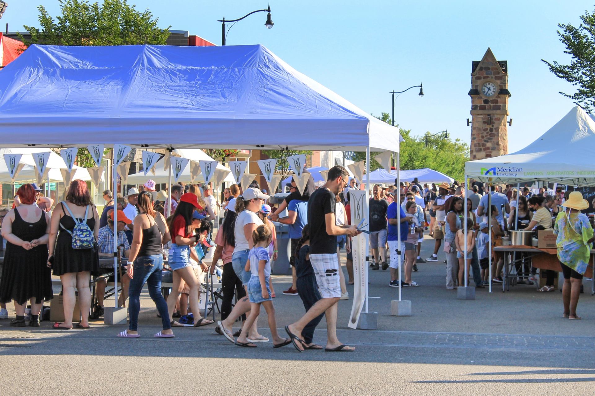 People attend a busy outdoor street fair with white and blue vendor tents and a stone clock tower in the background.