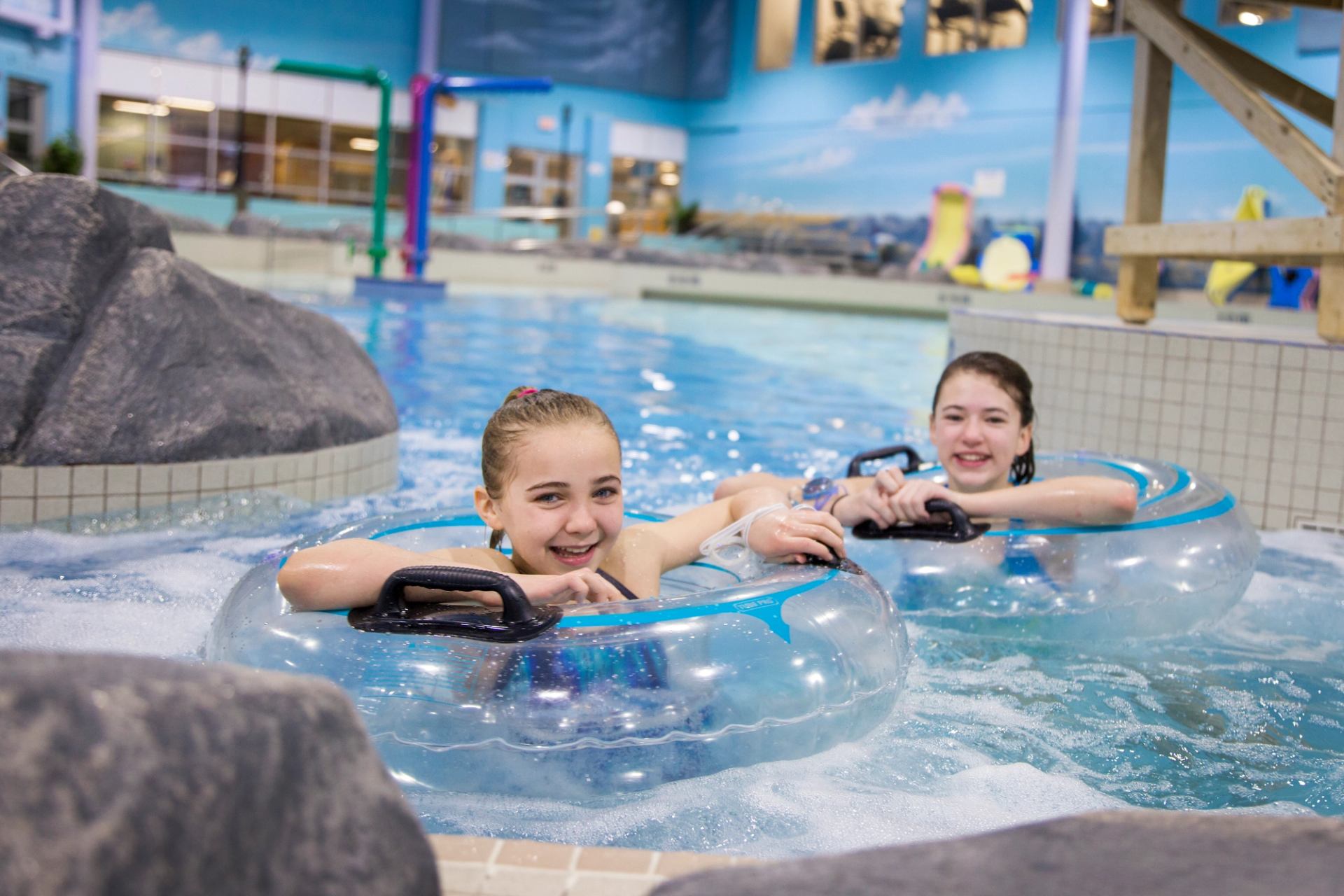 Indoor pool, two people ride blue inner tubes over water next to a slide wall.
