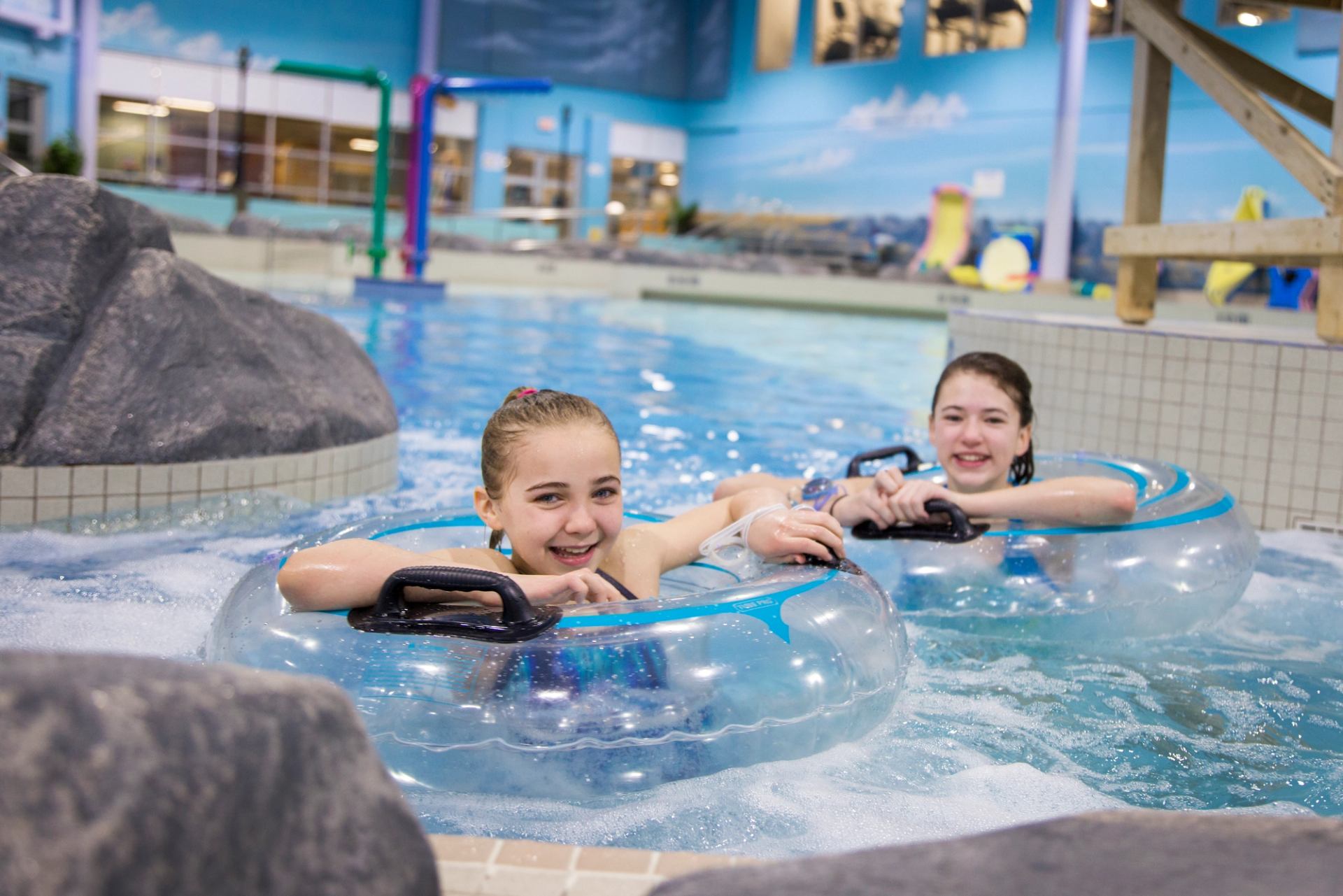 Indoor pool, two people ride blue inner tubes over water next to a slide wall.
