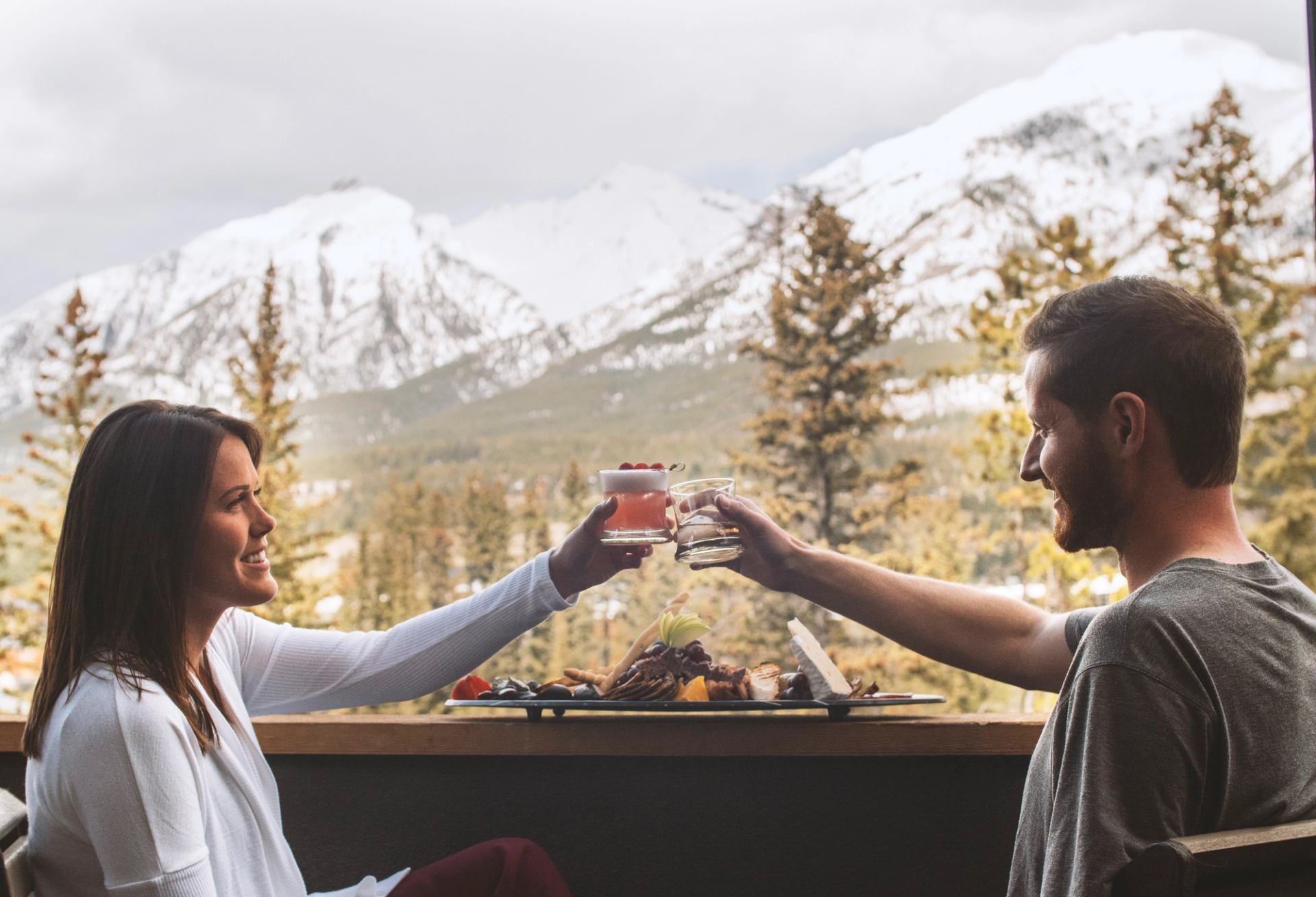 Two people toast drinks over a meal with snow-covered mountains behind them.