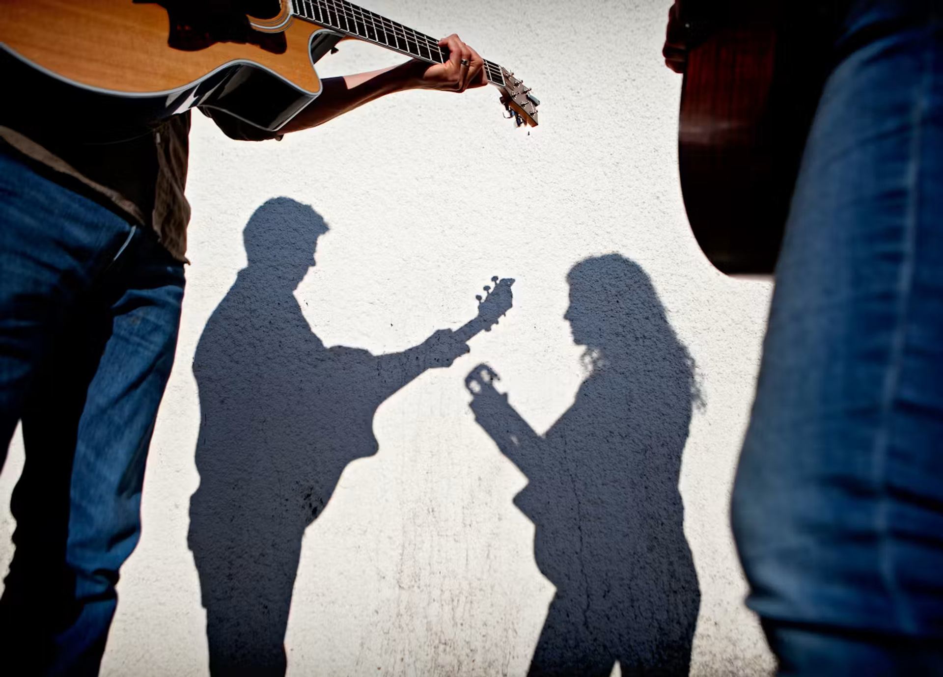 Shadows of two people playing guitars on a white wall, framed by musicians’ legs.