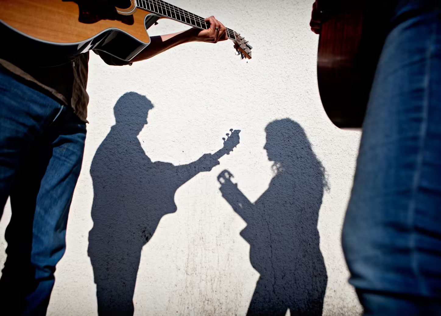 Shadows of two people playing guitars on a white wall, framed by musicians’ legs.