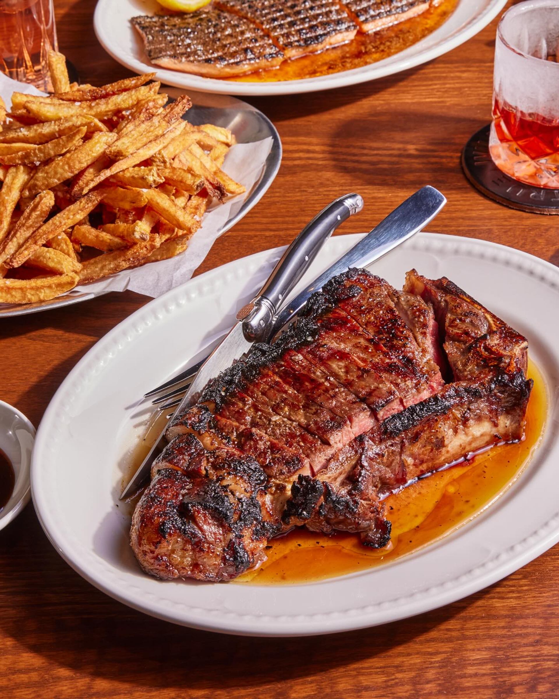 Grilled steak served with golden fries on a white plate, with cocktails and shared dishes on a wooden table.