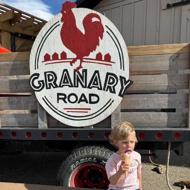 Sign reading “Granary Road” on a wooden fair booth, with a child holding an ice cream.