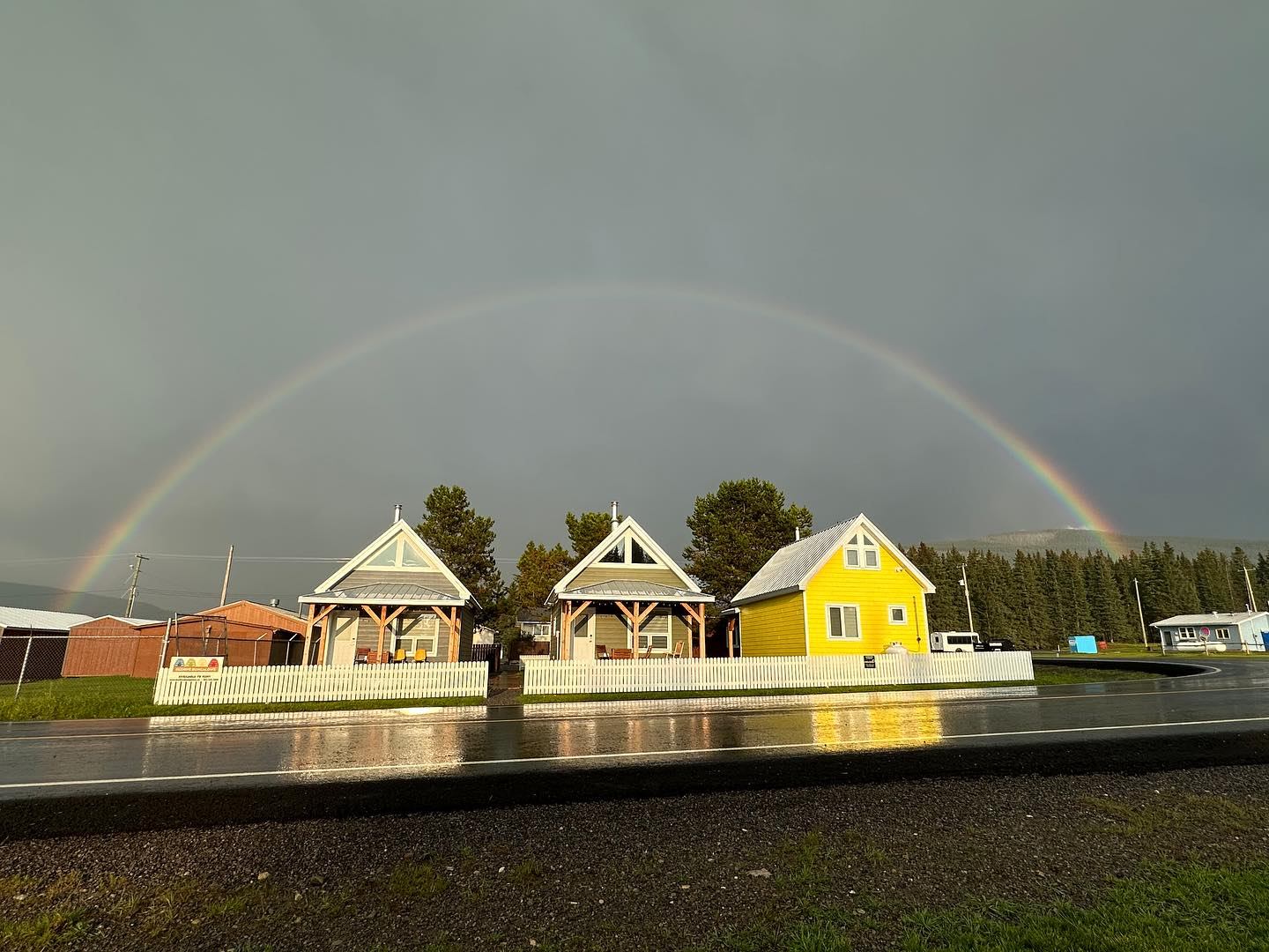 Colorful bungalows with a full rainbow overhead on a wet, reflective roadway.