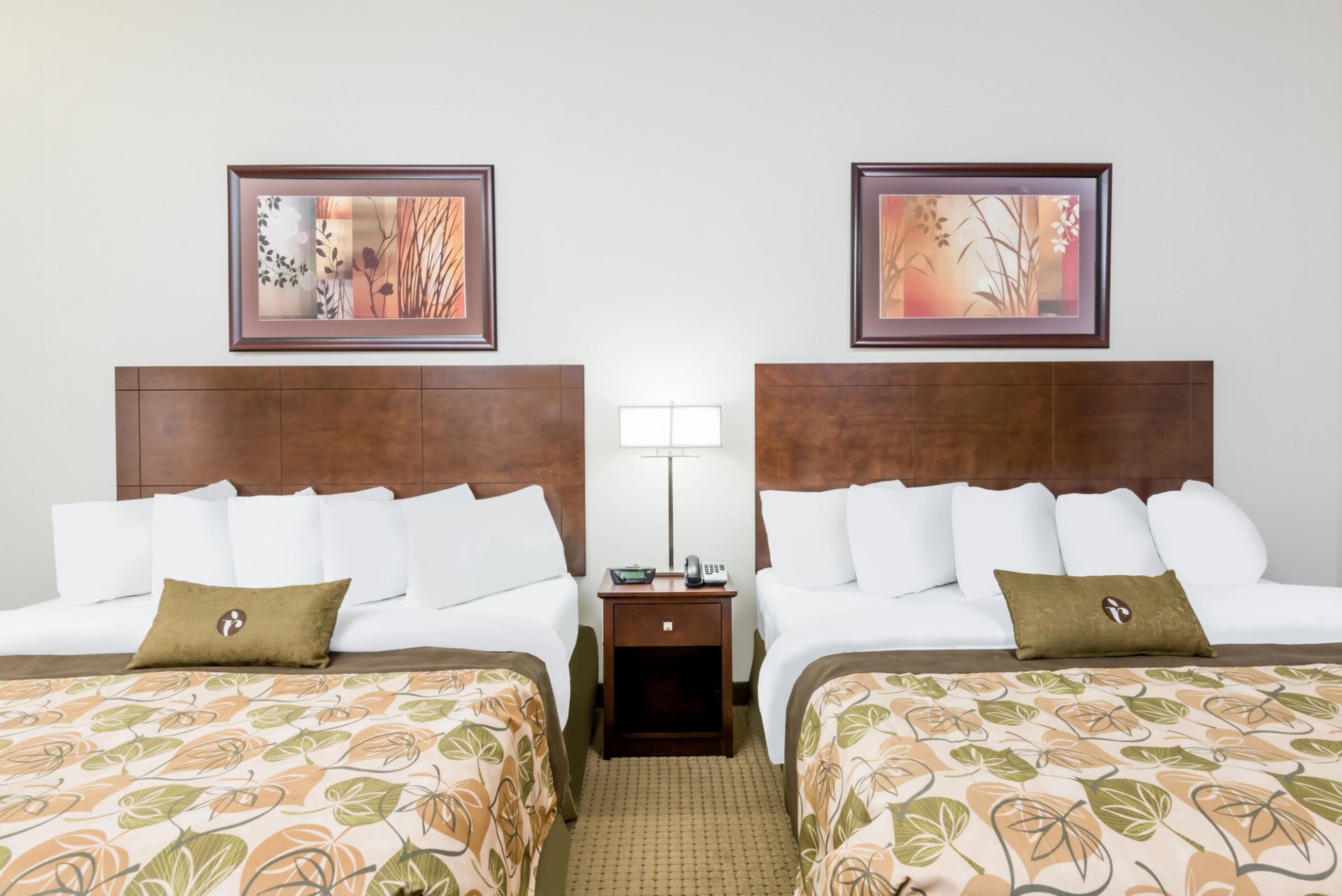 Hotel room with two double beds, floral bedspreads, bedside table and wall art.