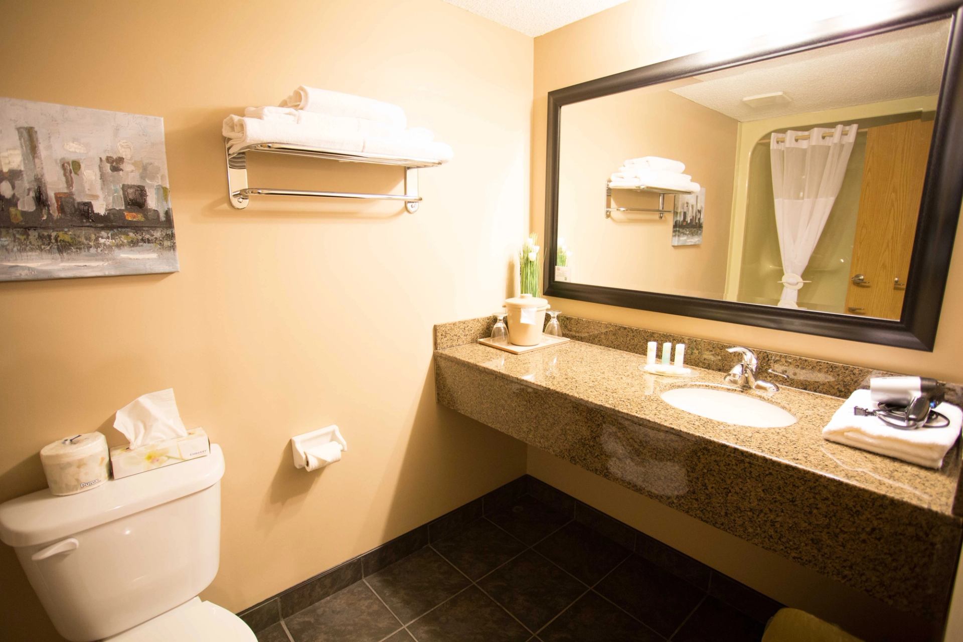 Modern bathroom with beige walls, dark tile floor, granite countertop, and large mirror. Includes a sink, toiletries, and a neatly folded towel.