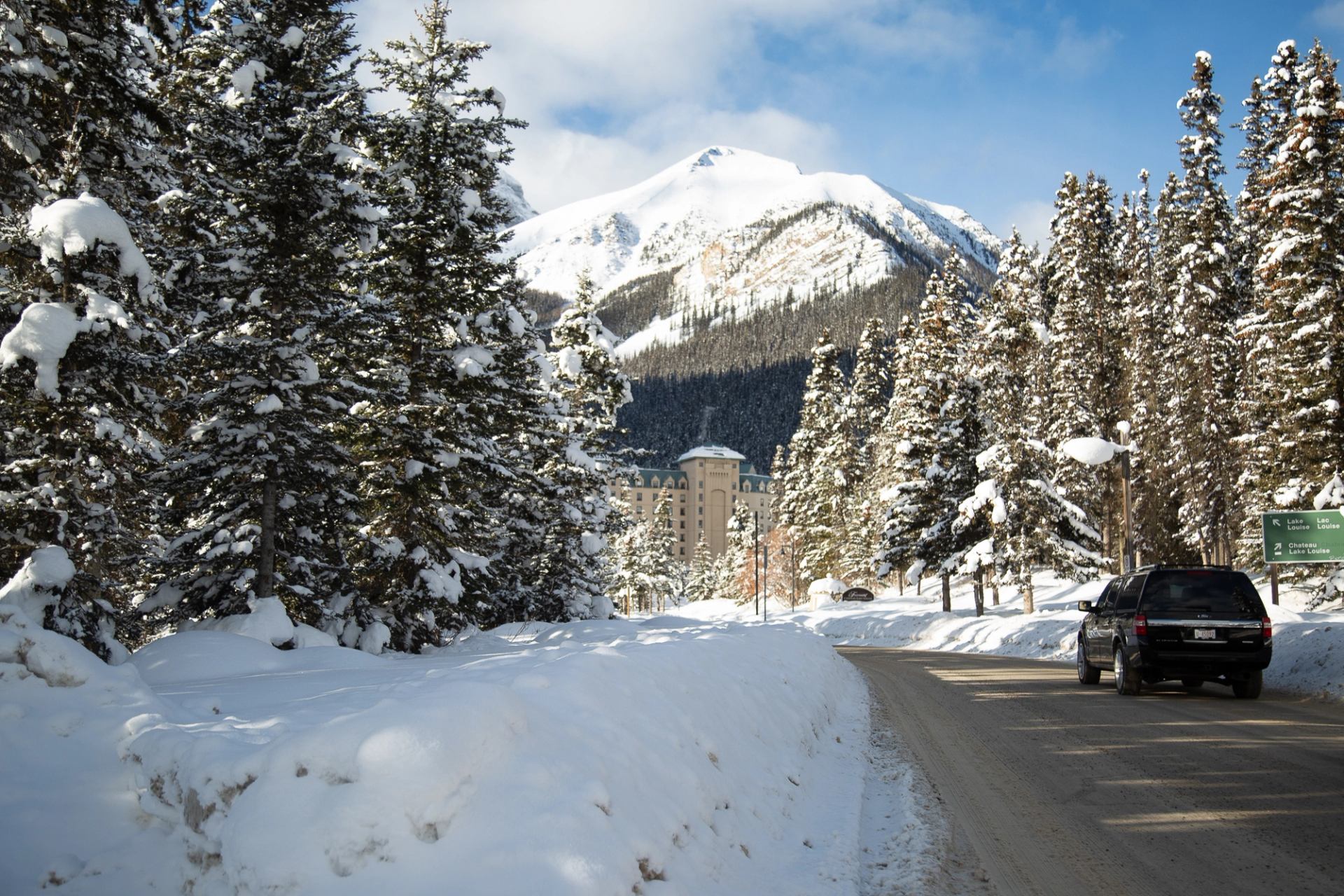 Black SUV traveling on a snow-covered road lined with pine trees and mountain peaks