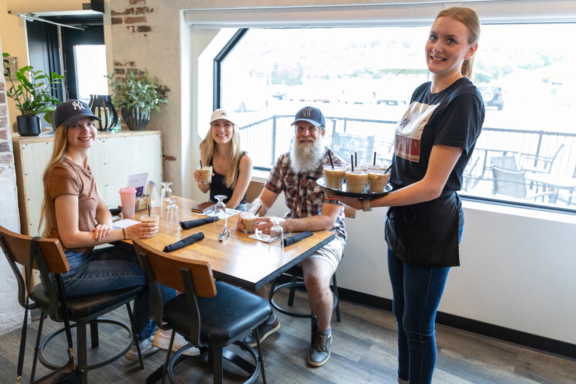 Server carrying iced coffees to three people sitting at a café table by window.