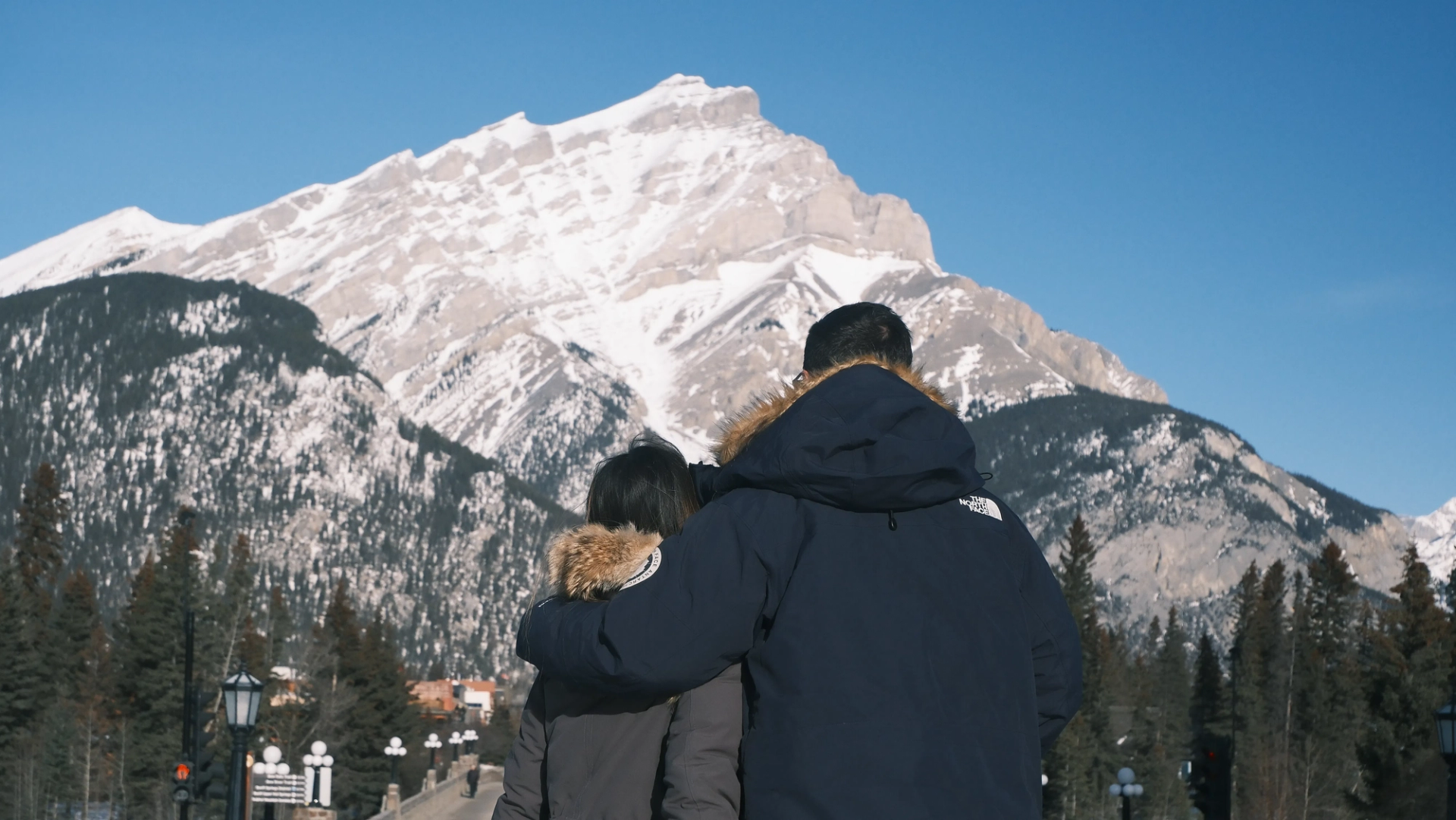 Two people in winter jackets stand on a bridge facing a snow-covered mountain.