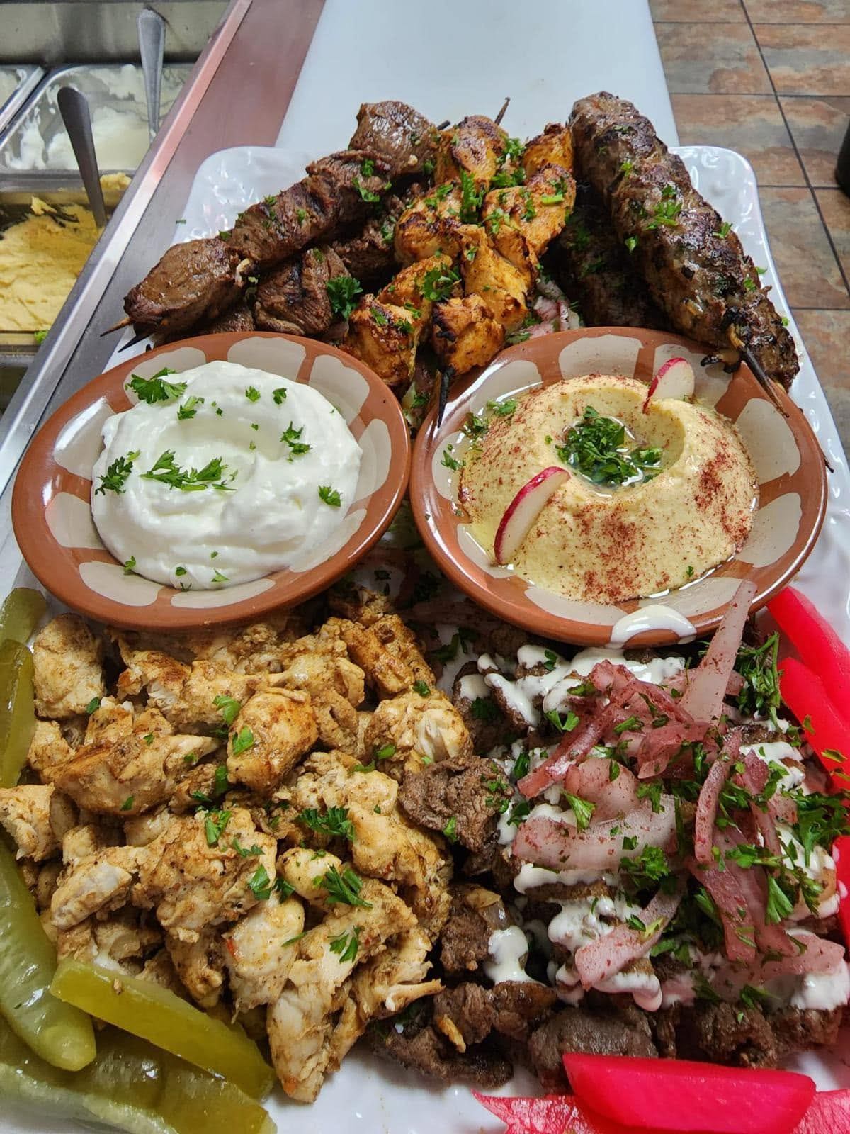 A platter showcases grilled meats, hummus, garlic sauce, pickles, and seasoned chicken pieces.