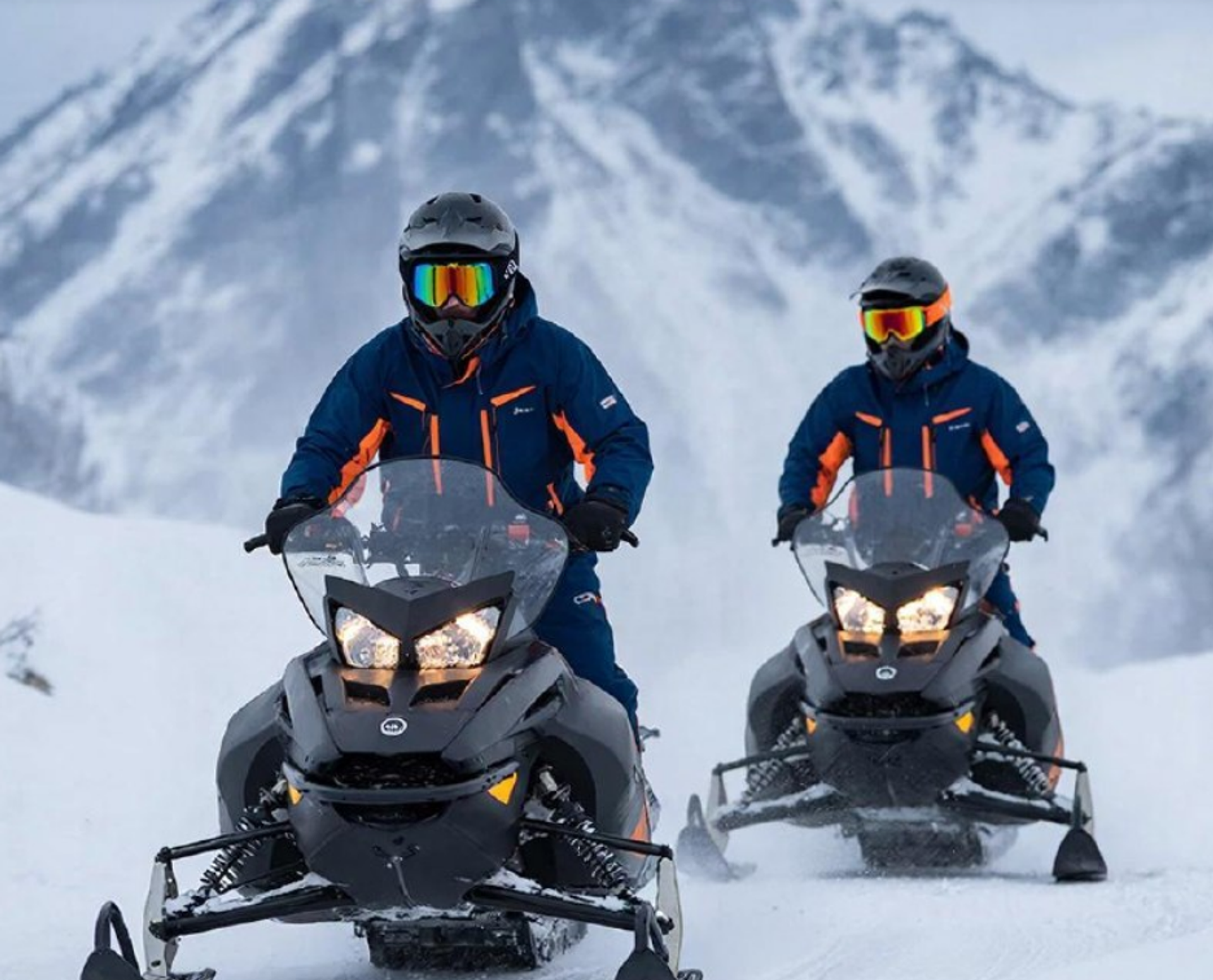 Snowmobilers ride across snow with rugged mountains in the background.