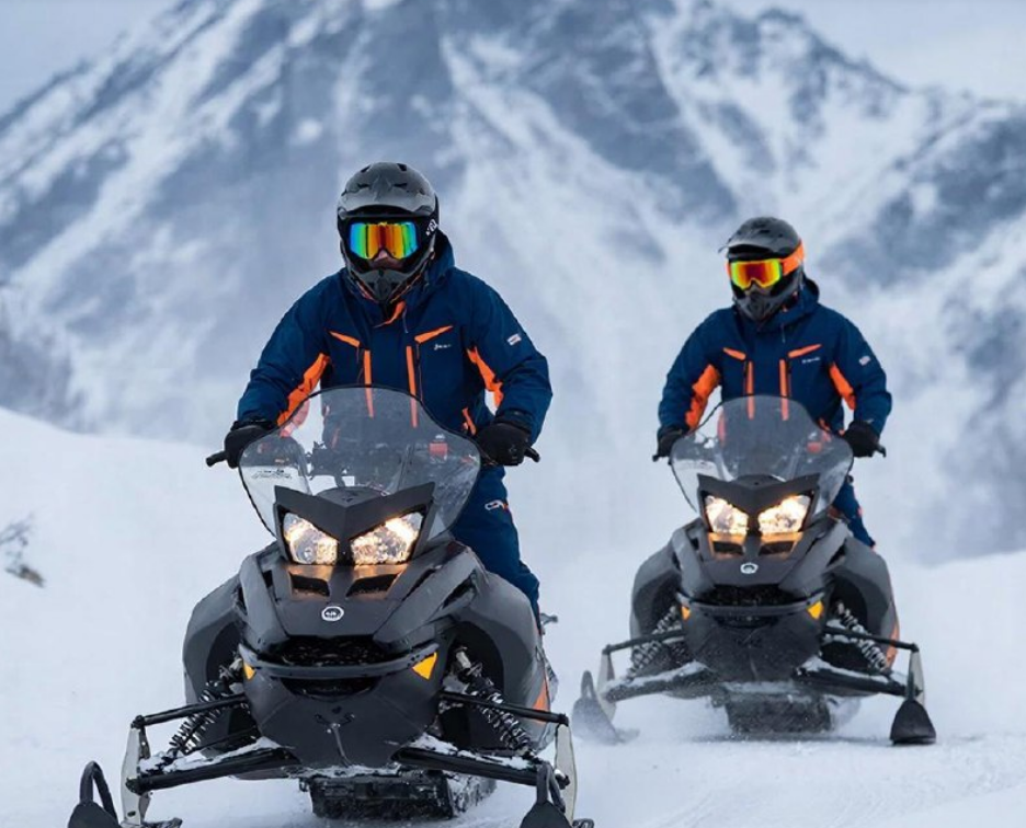 Snowmobilers ride across snow with rugged mountains in the background.