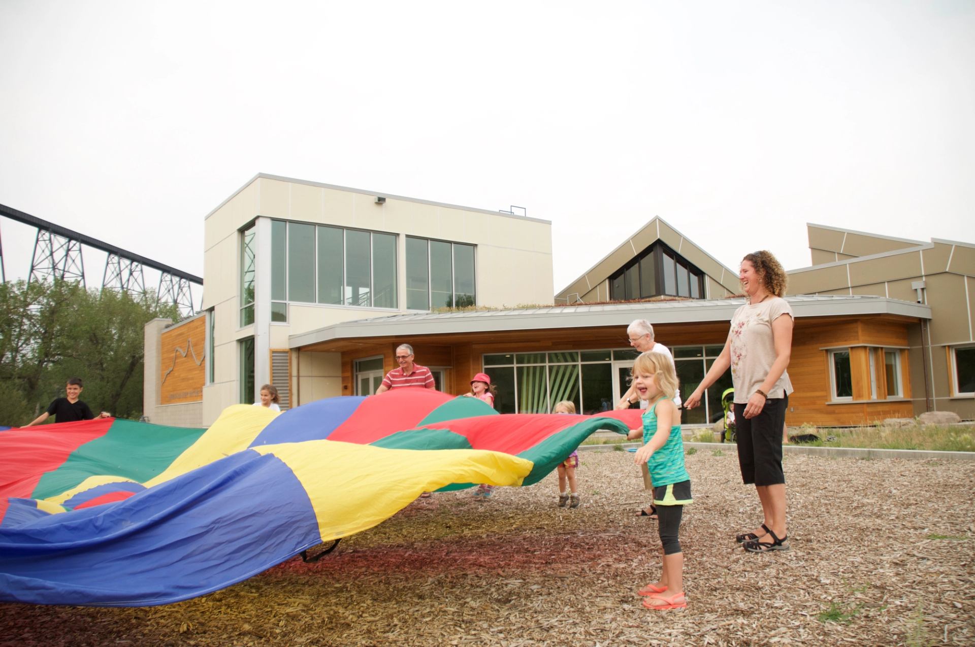 Children and adults lift a bright parachute together in front of a nature centre.