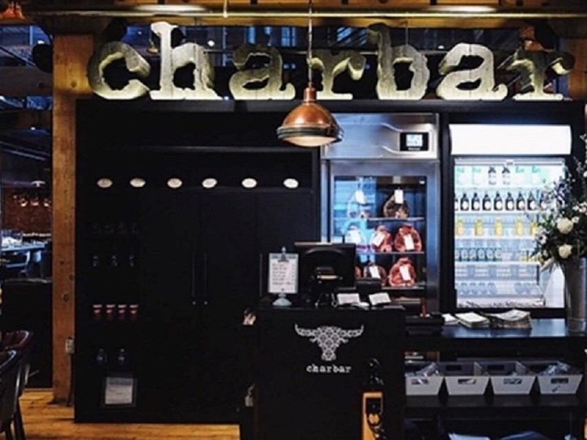 Modern bar interior with a large illuminated "charbar" sign above. Shelves with drinks and decorative elements create a cozy, inviting atmosphere.