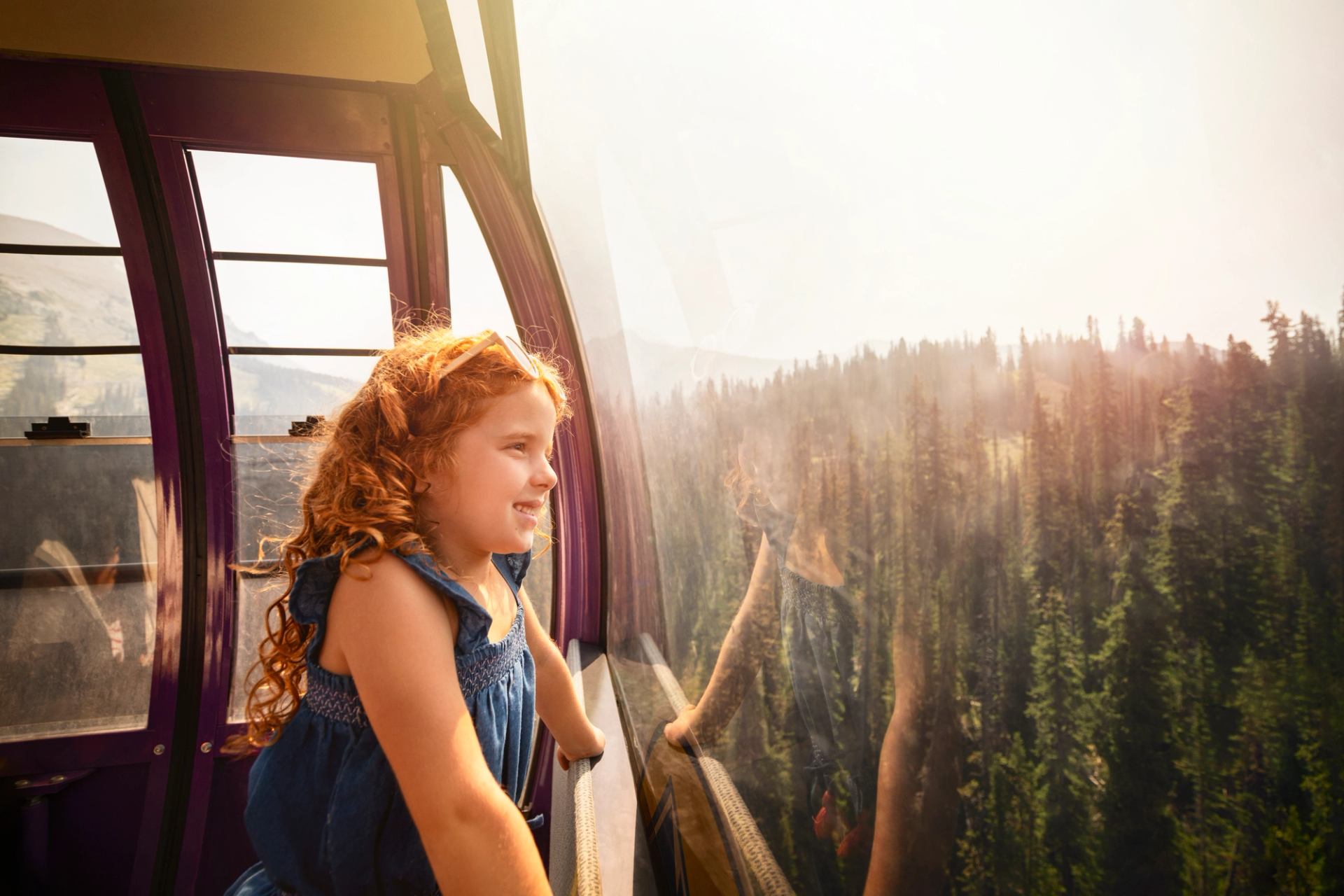 Child in gondola cabin looks out window at forested mountain landscape below.