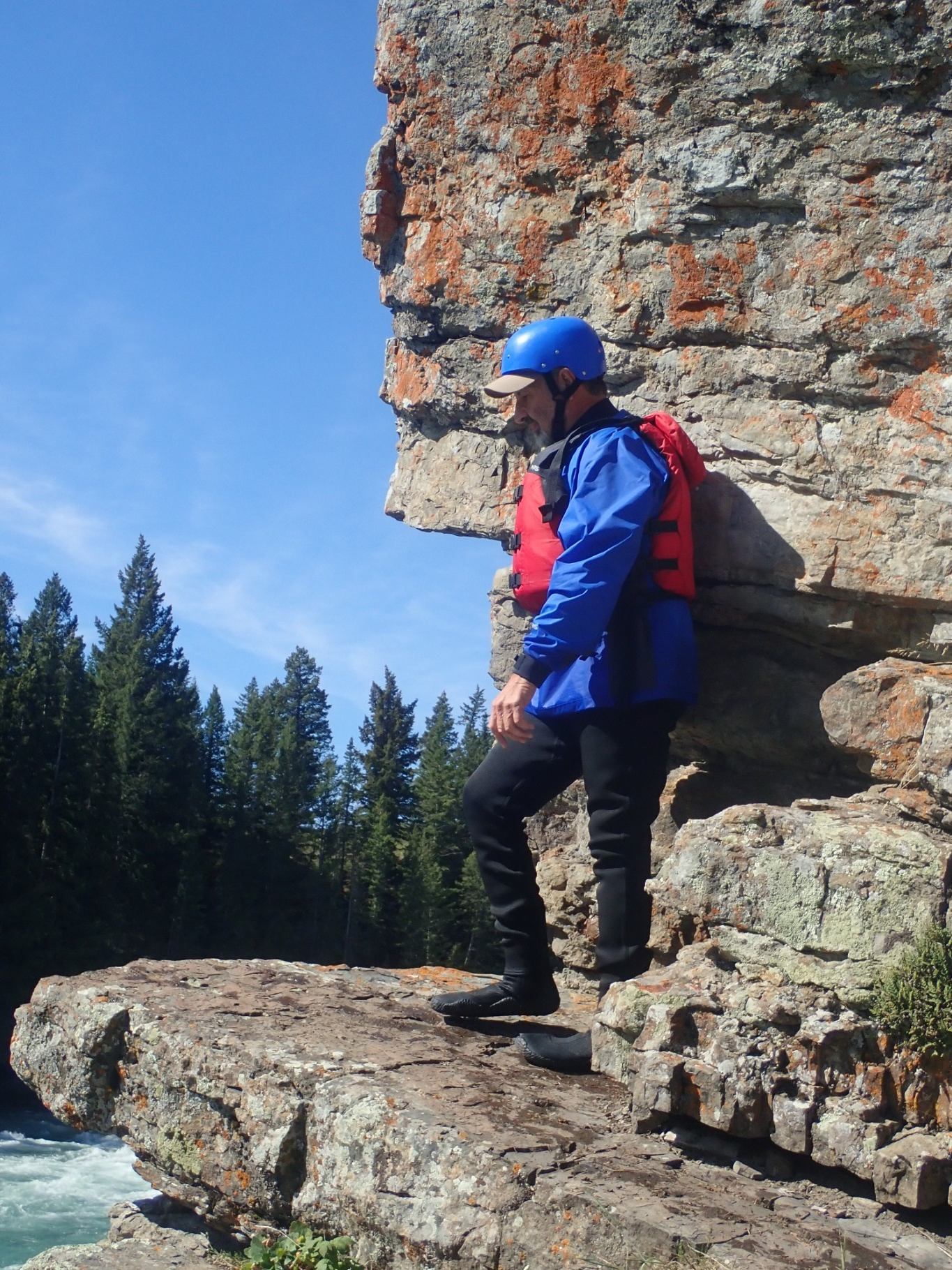 Person in helmet and life jacket stands on rocky cliff above river and forest.