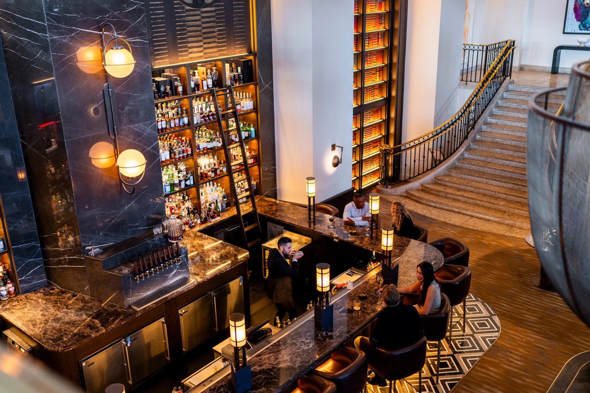 Modern bar with marble counters, tall shelves of bottles, and a sweeping staircase.