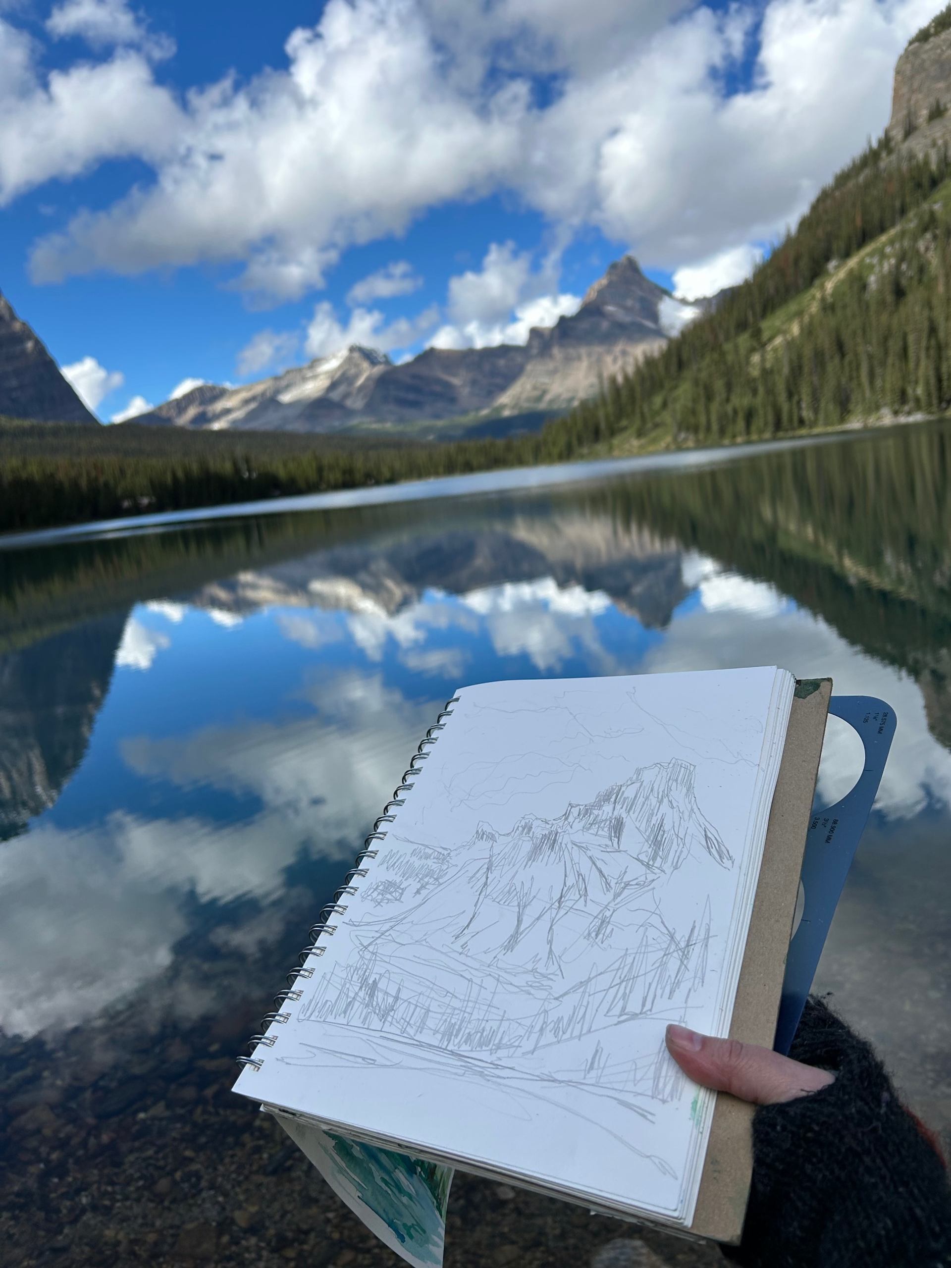 A person holding a sketch book with the lake and mountains in the background.