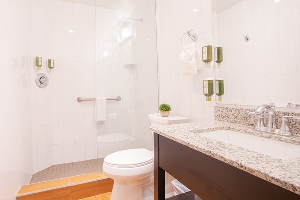 A bathroom with white tile walls features a walk-in glass shower with grab bars and a toilet beside a granite countertop sink. Mounted toiletry dispensers and bright lighting create a clean, accessible space.