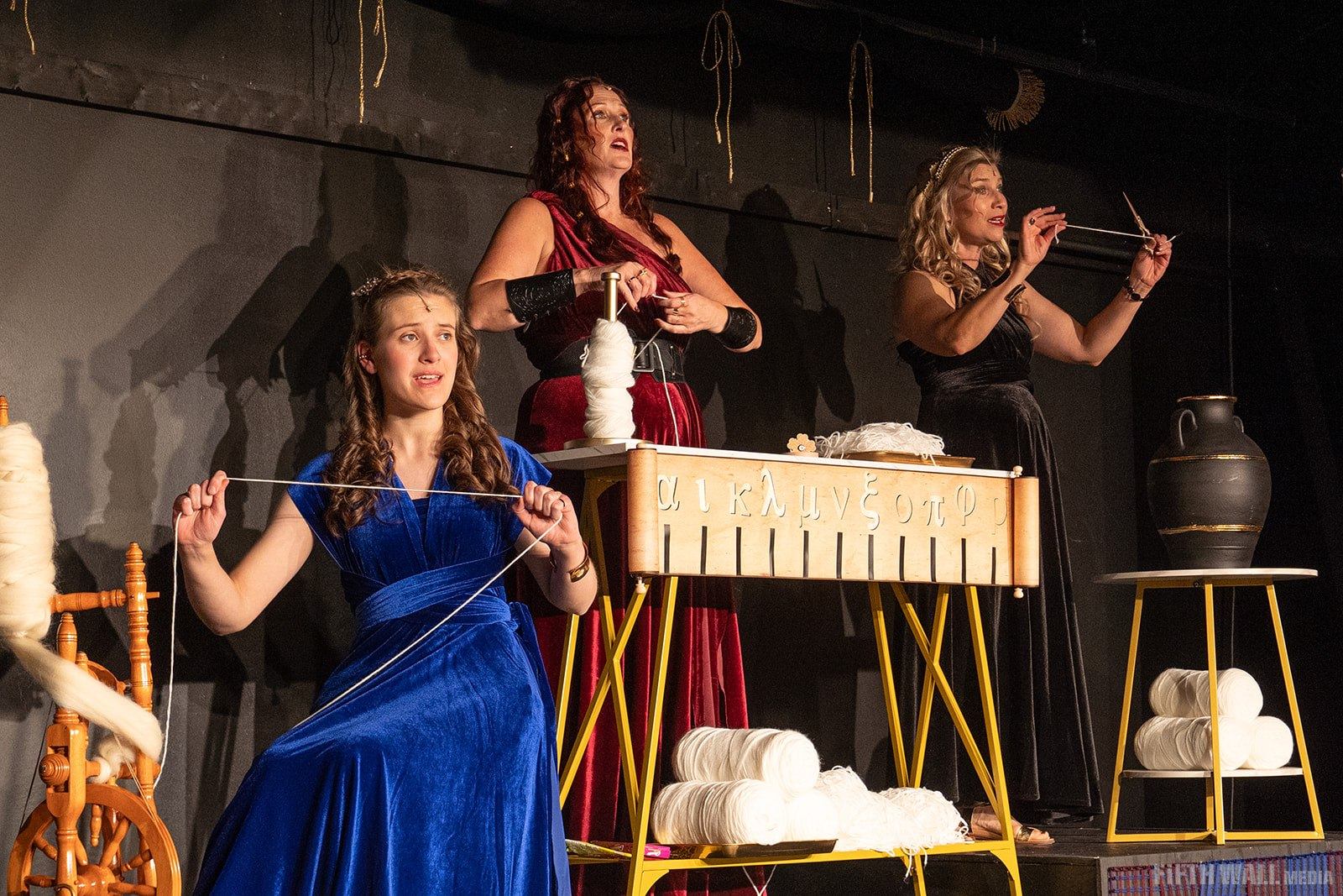 Three performers in long dresses spin and measure yarn onstage beside props and spindles.